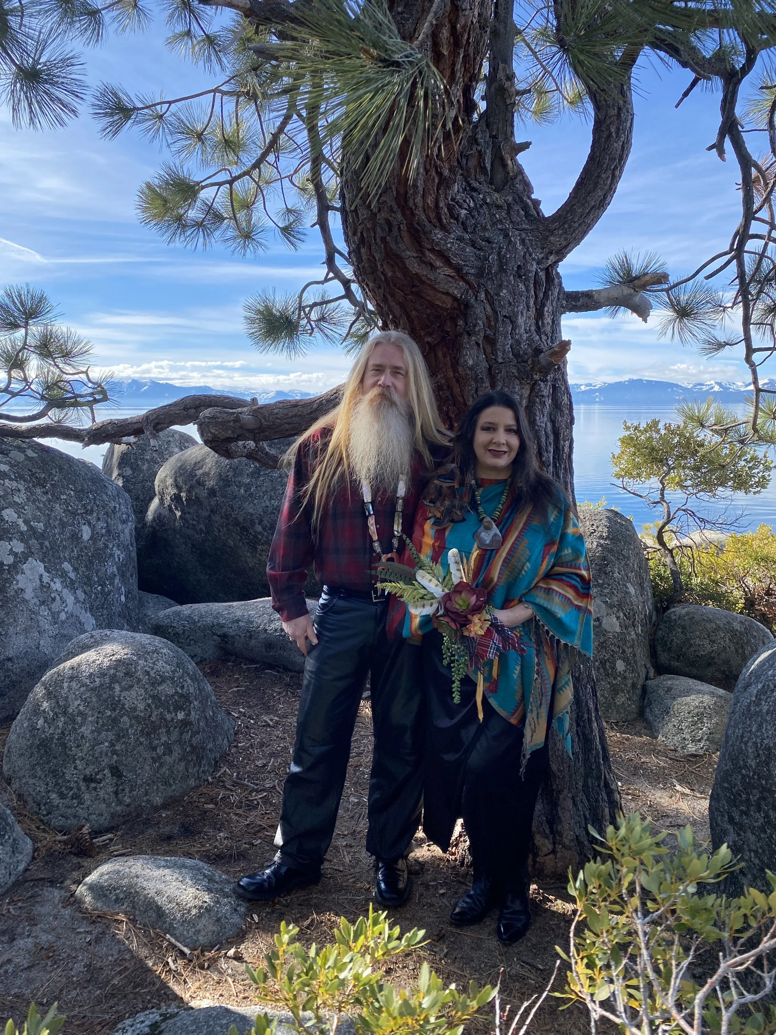 A bride dressed in a colorful poncho holding a bouquet with her groom, standing in front of a large pine tree on a rocky hillside overlooking a lake and snow-capped mountains on a sunny day at Sand Harbor after their spiritual ceremony.