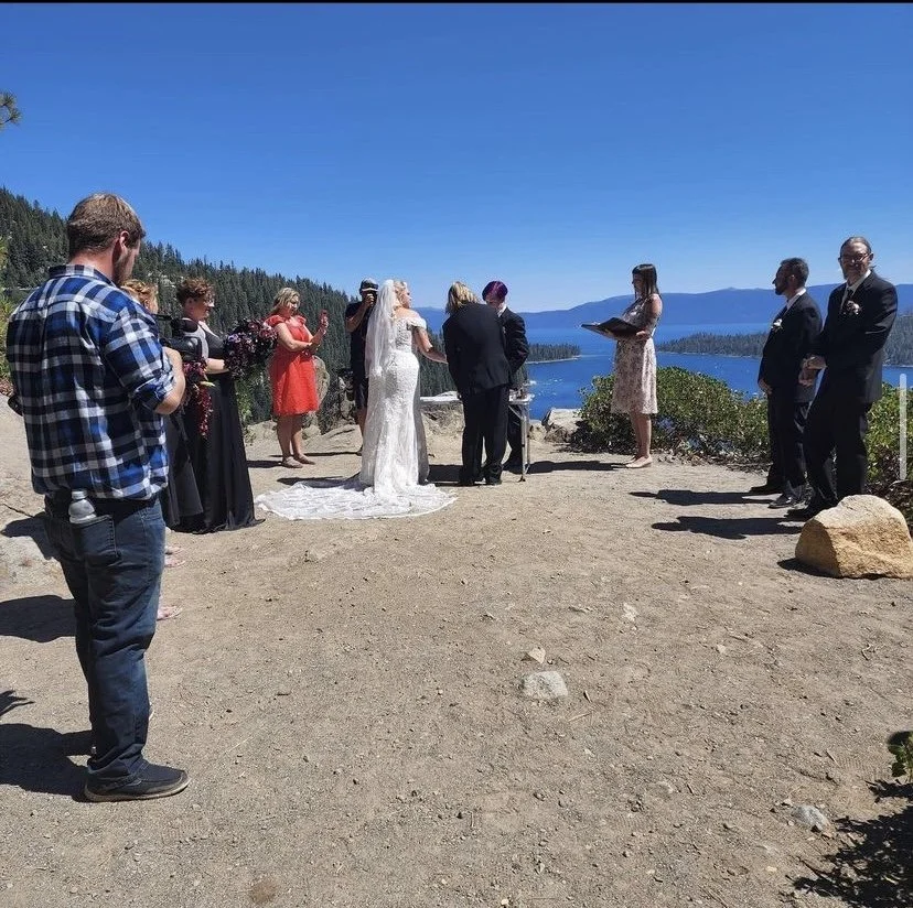 A wedding ceremony taking place outdoors on a sunny day near a body of water, with the bride and groom exchanging vows. The bride is in a white dress with a long veil, and the groom is in a dark suit. Bridesmaids and groomsmen stand on either side, w
