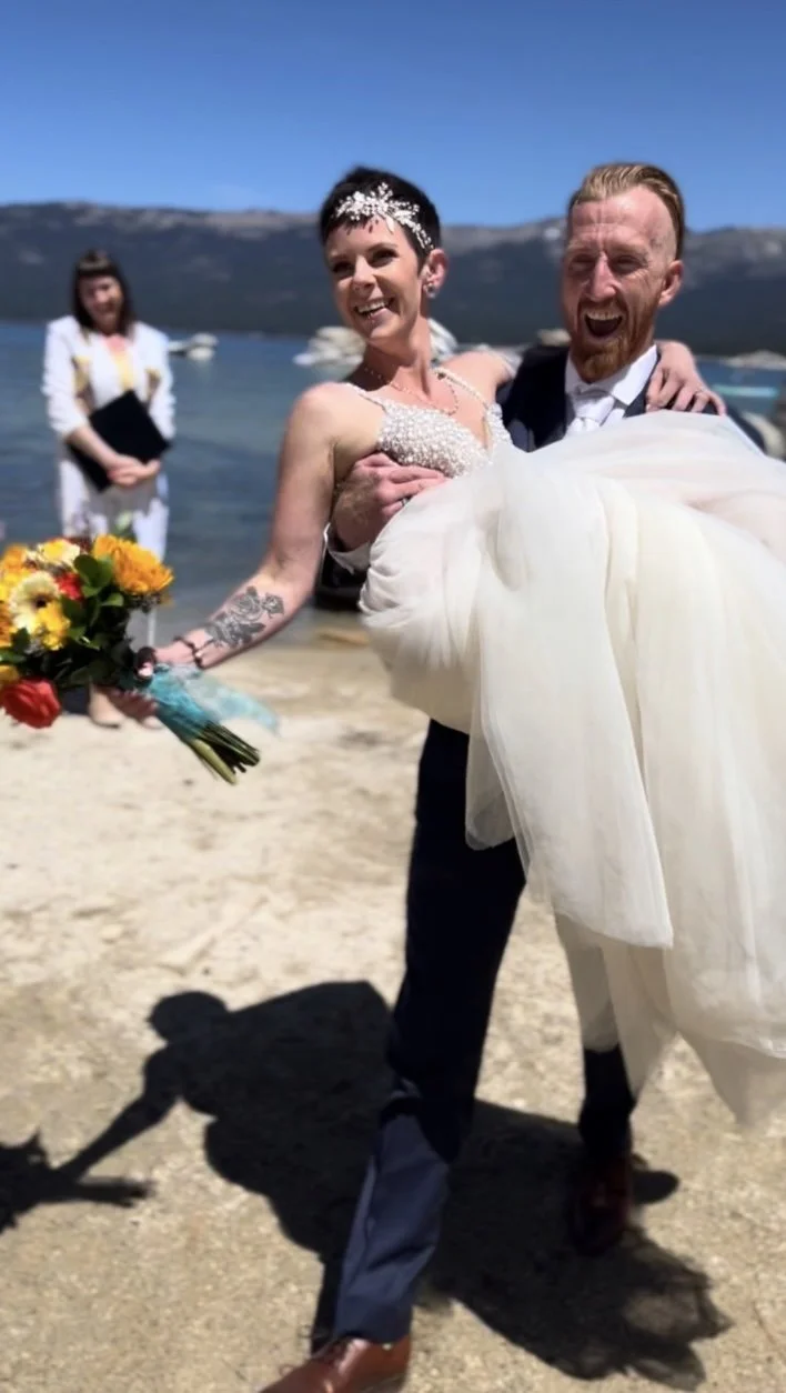 A happy couple in wedding attire, with the groom carrying the bride, standing on a beach with water and mountains in the background, during a sunny day.