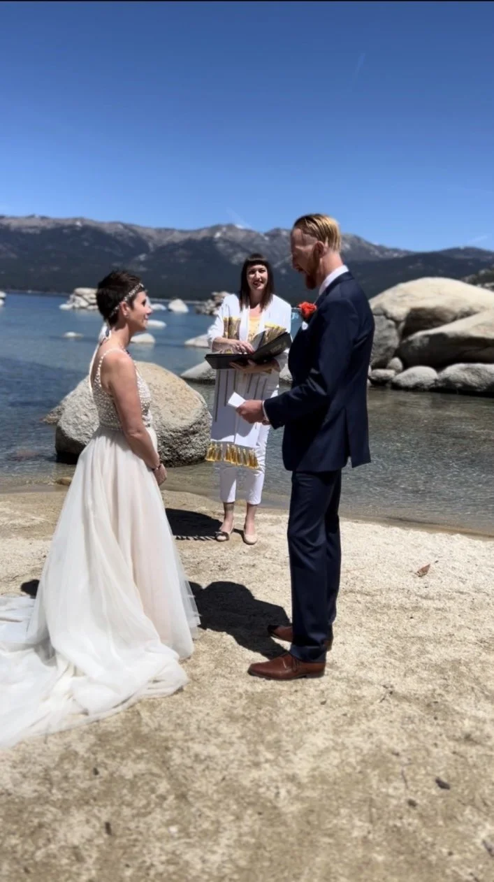 A couple getting married on a sandy beach by a lake, with mountains in the background. The bride is in a white gown, and the groom is in a dark suit. An officiant stands behind them, holding a book.