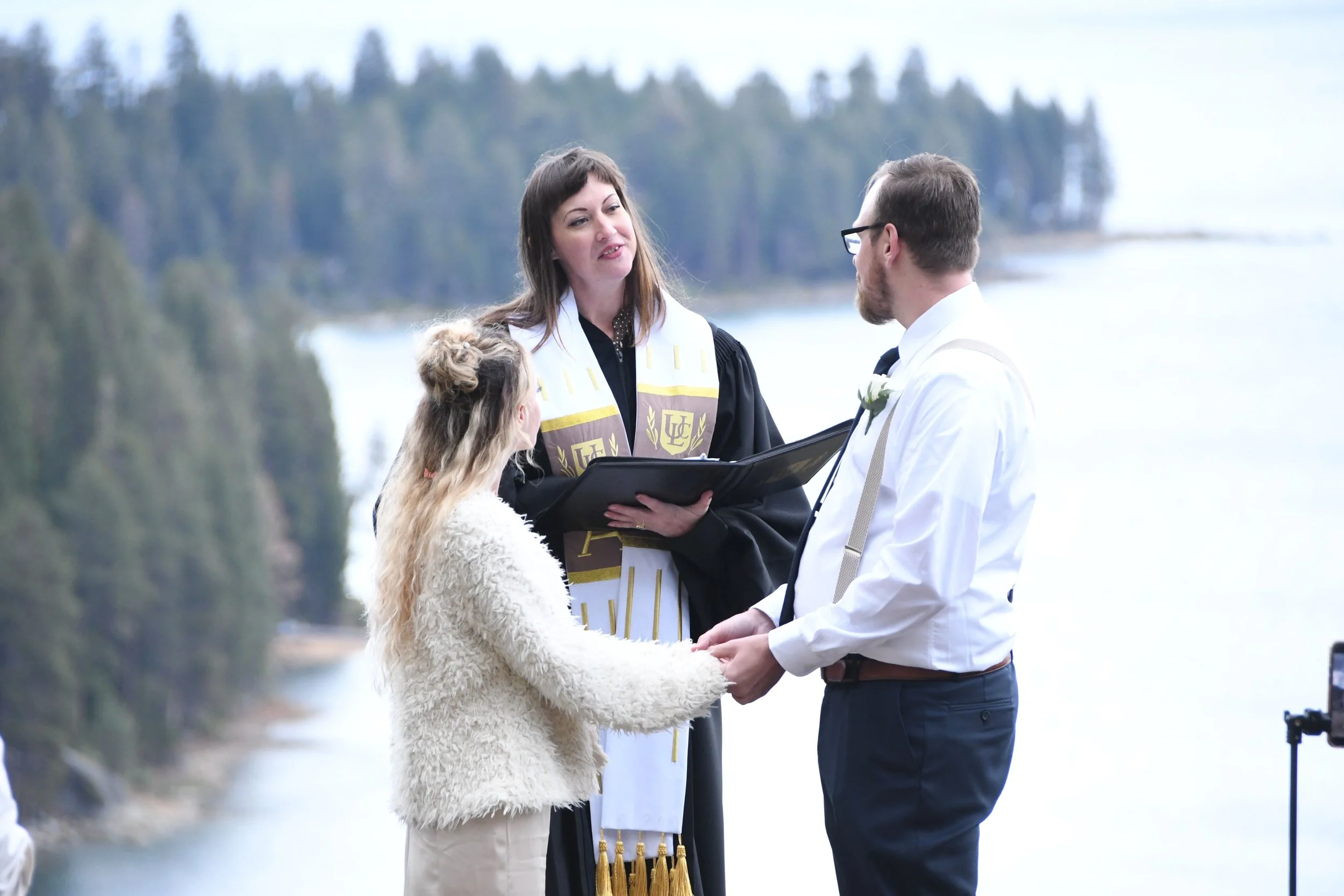 A couple gets married outdoors by a river, with an officiant officiating, while a young girl holds their hands during the ceremony.