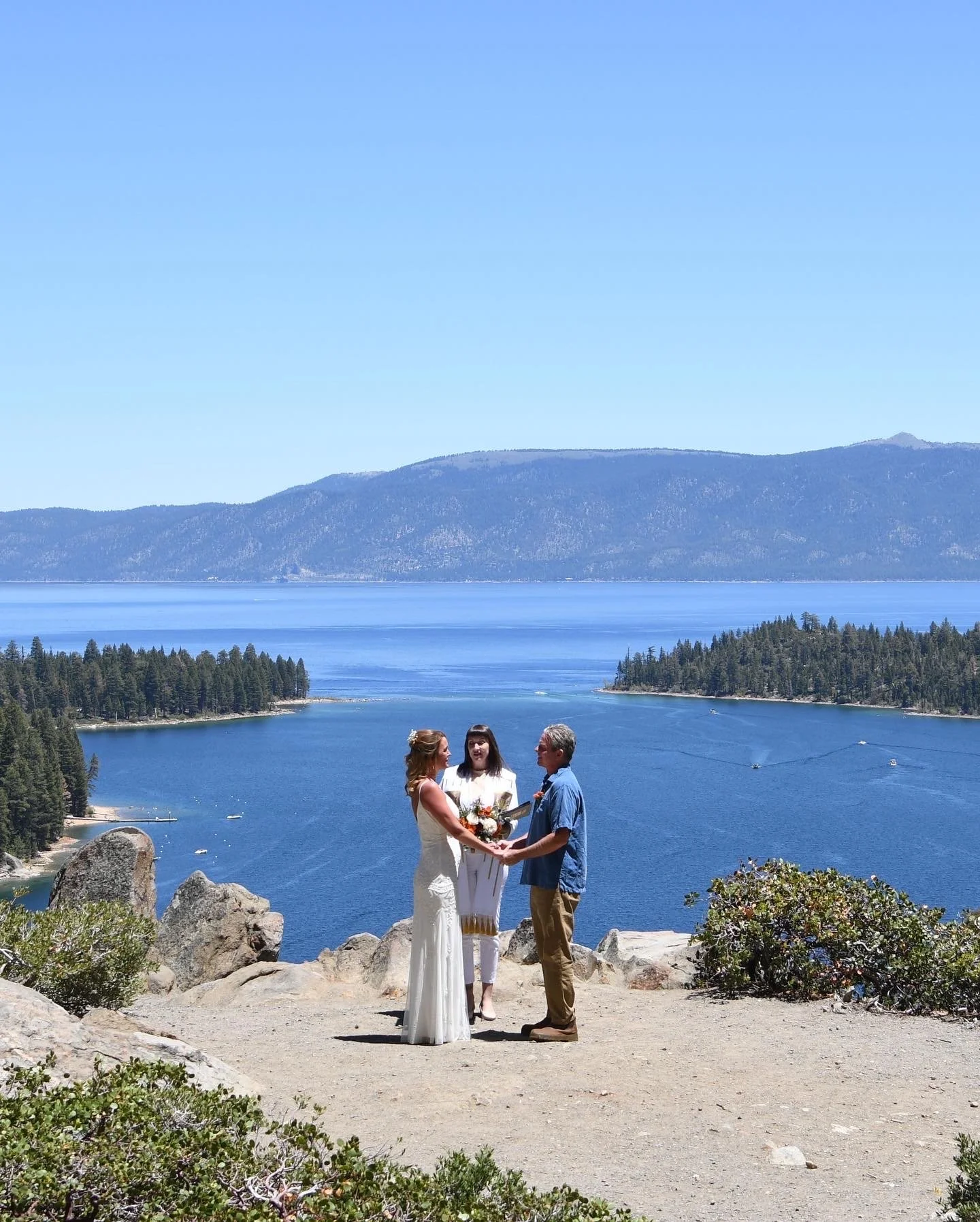 A wedding ceremony taking place outdoors on a rocky ledge with a scenic view of Emerald Bay, forested islands, and mountains in the background. The bride and groom are holding hands and facing each other, with an officiant standing behind them.