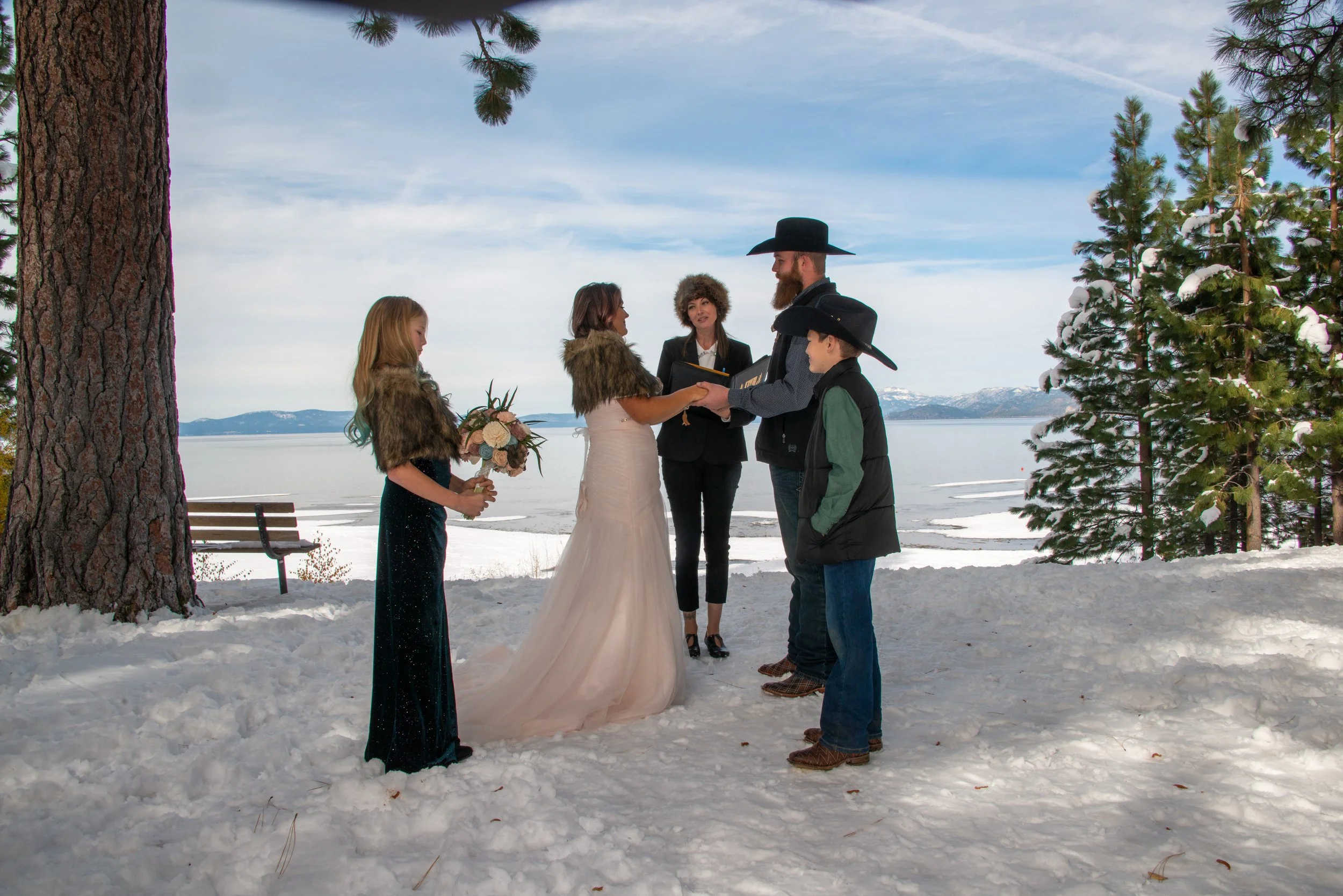 A wedding ceremony taking place outdoors in a snowy landscape by a lake, with a bride, groom, two children, and an officiant, surrounded by pine trees and mountains in the background.