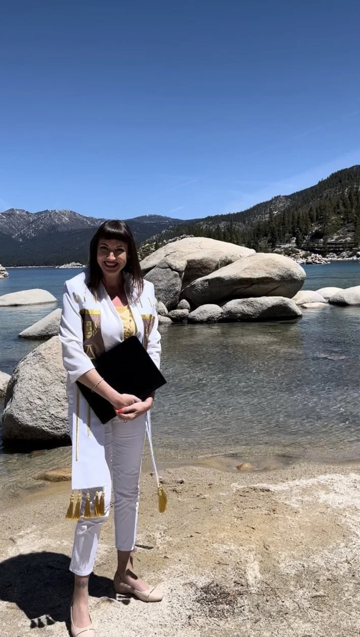 Officiant in a white suit standing on a sandy beach at Sand Harbor by a lake, with large rocks and forested mountains in the background, under a clear blue sky.