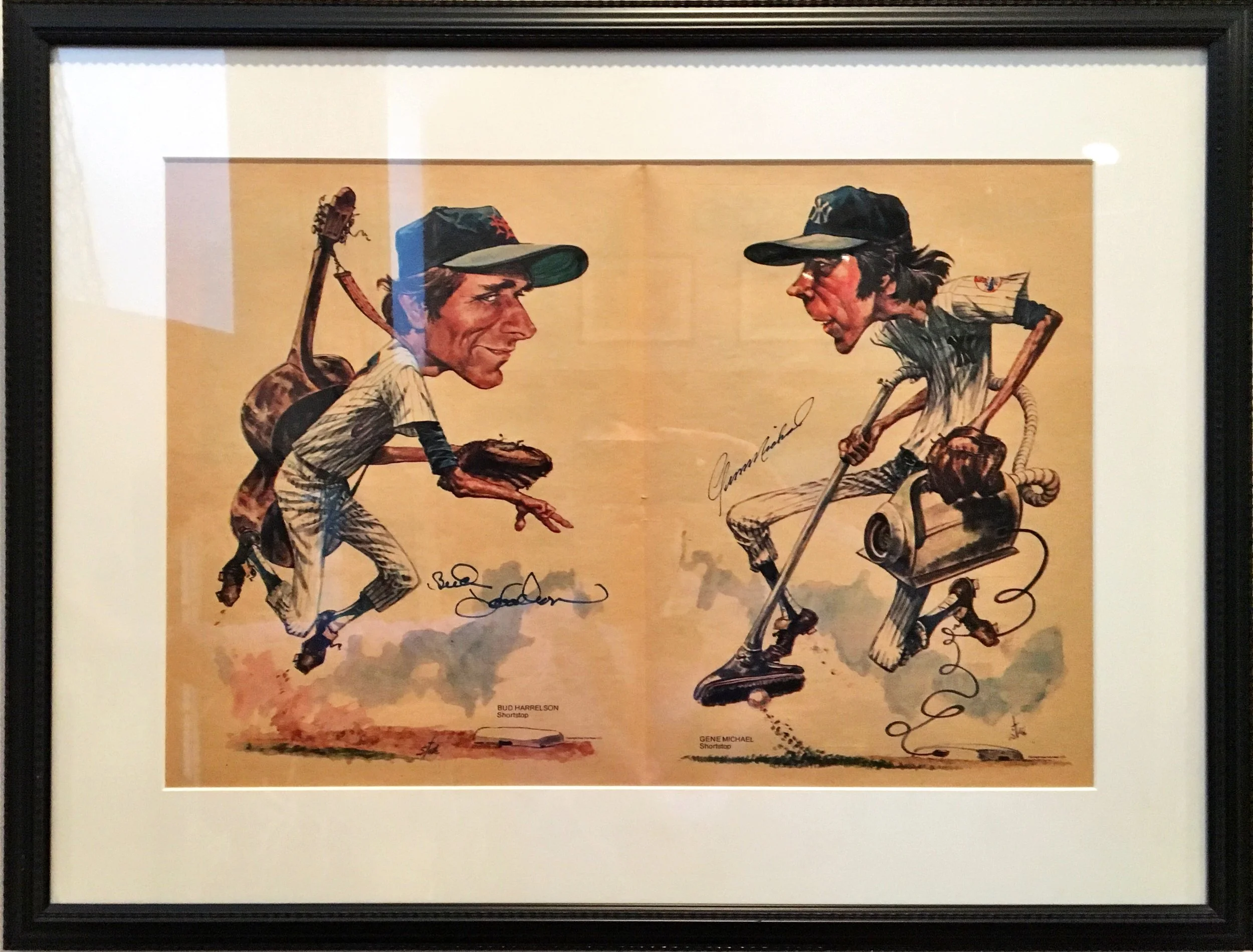 A framed caricature illustration of two baseball players, one with a guitar on his back and the other with a cane, both dressed in striped tops and caps, engaging in a playful exchange. Signed signatures are at the bottom, with a signature on the left labeled 'SHUD HARRELSON Shortstop' and on the right 'GENE MICHAEL Shortstop'.
