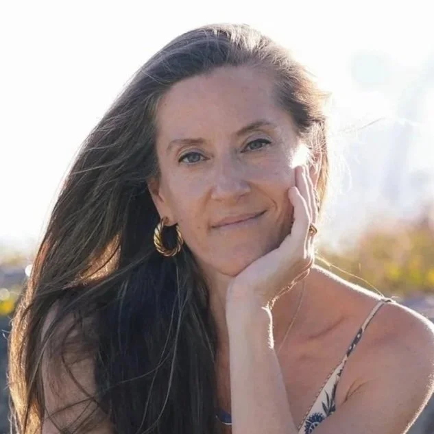 A woman with long brown hair, wearing gold hoop earrings and a sleeveless patterned top, smiling softly while resting her face on her hand outdoors against a bright background.