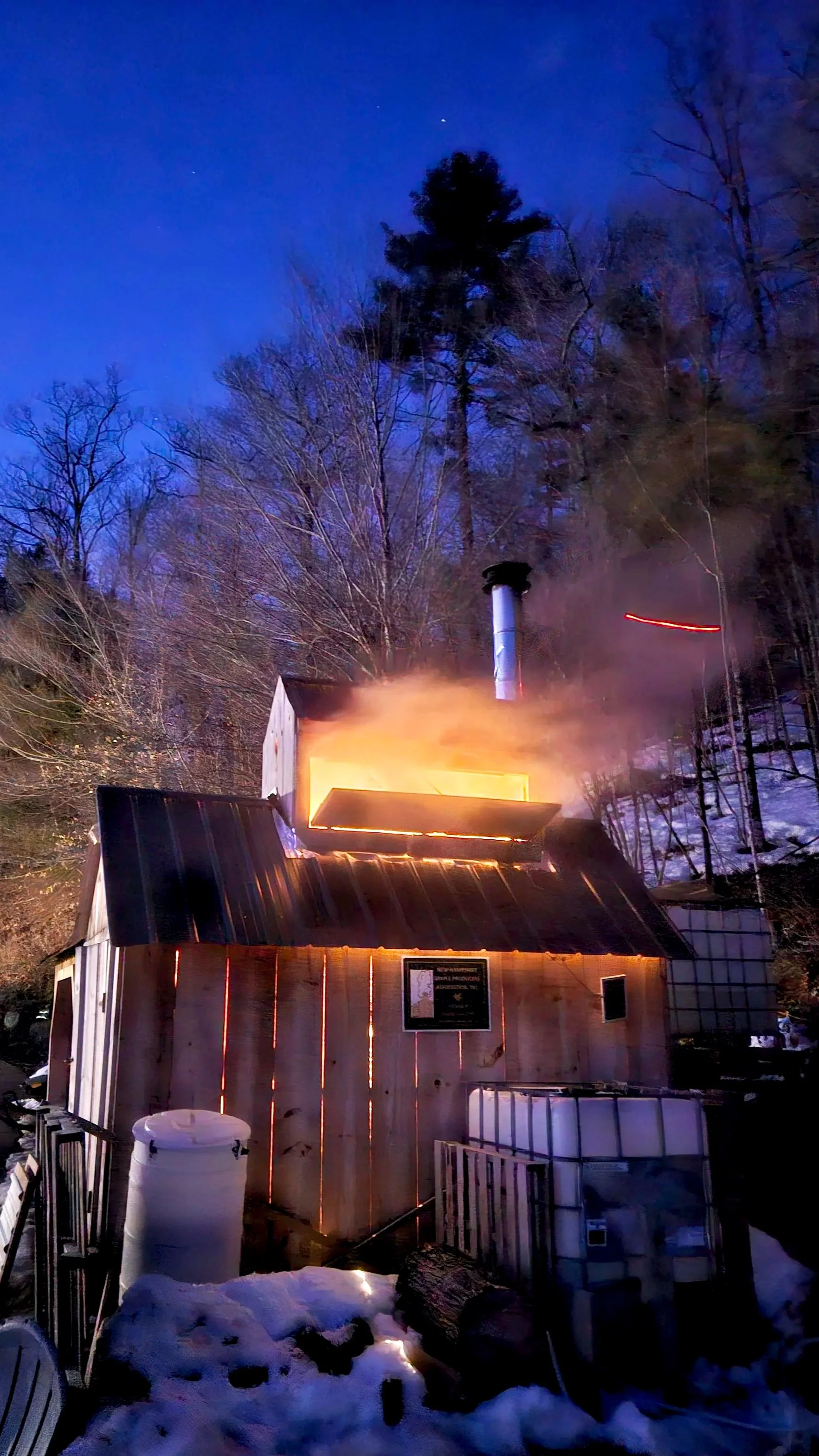 A wooden barn producing maple syrup, with steam and smoke emanating from the chimneys, surrounded by snow and trees during dusk or night.
