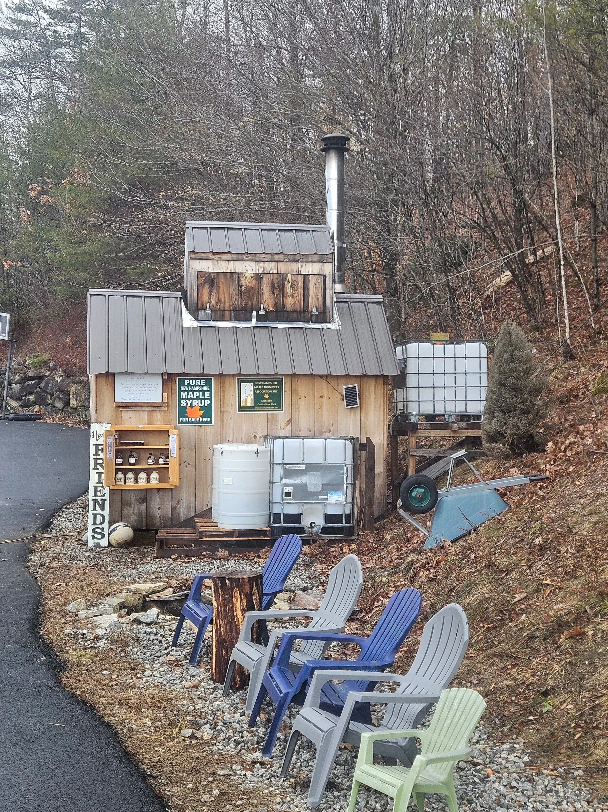 A small wooden building with signs and a chimney, surrounded by outdoor chairs, containers, and tools on a gravel area at the side of a road, with a hill and bushes in the background.