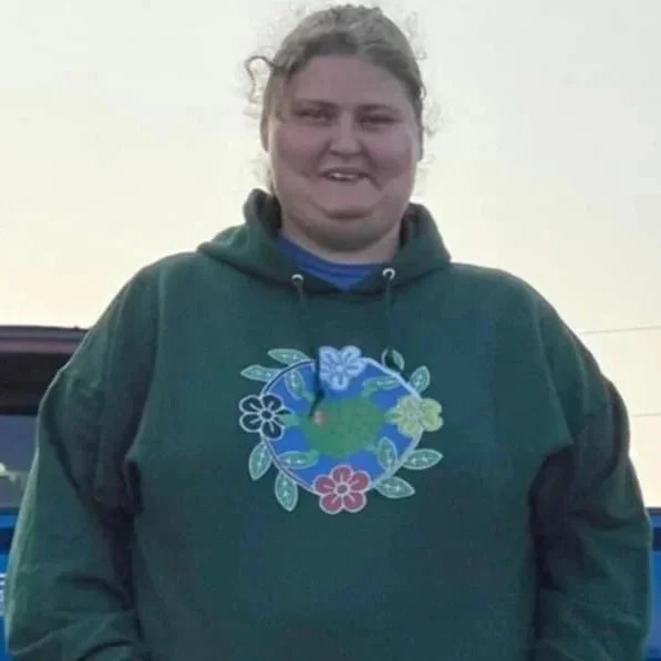 A woman standing outdoors wearing a dark green hoodie with a colorful Earth and floral design on the front.