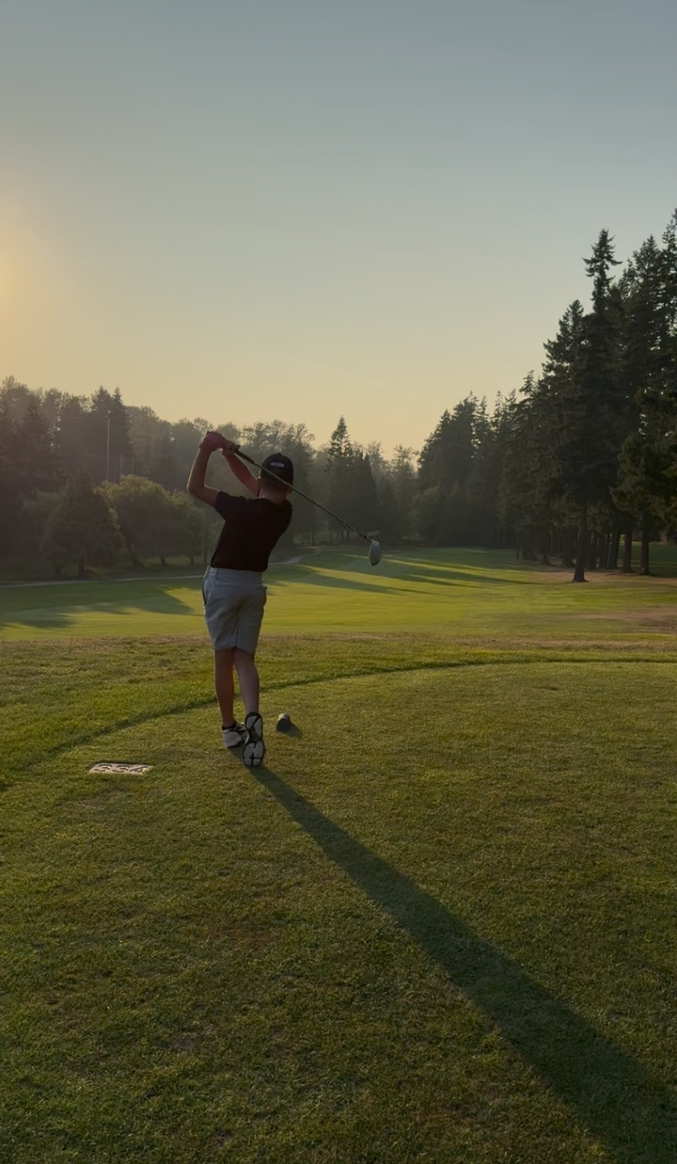 A person swinging a golf club on a golf course during sunset, with trees in the background.