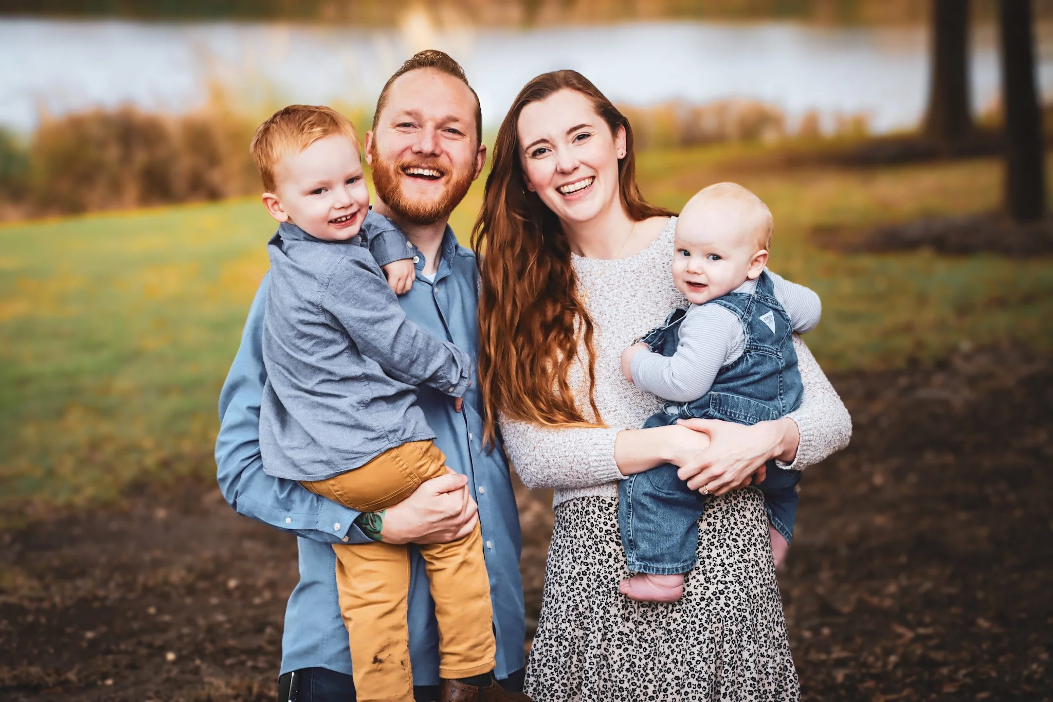 A happy family of four outdoors on a fall day, smiling at the camera. The father, with a beard, is holding a young boy with red hair and braces. The mother, with long red hair, is holding a baby girl with light hair and dressed in denim. The background shows a park with trees and a body of water.