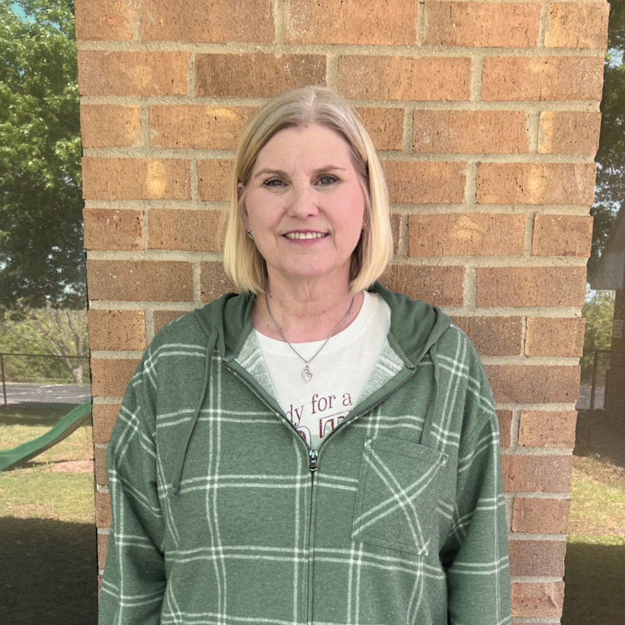 Portrait of a middle-aged woman with shoulder-length blonde hair standing against a brick wall outdoor background.