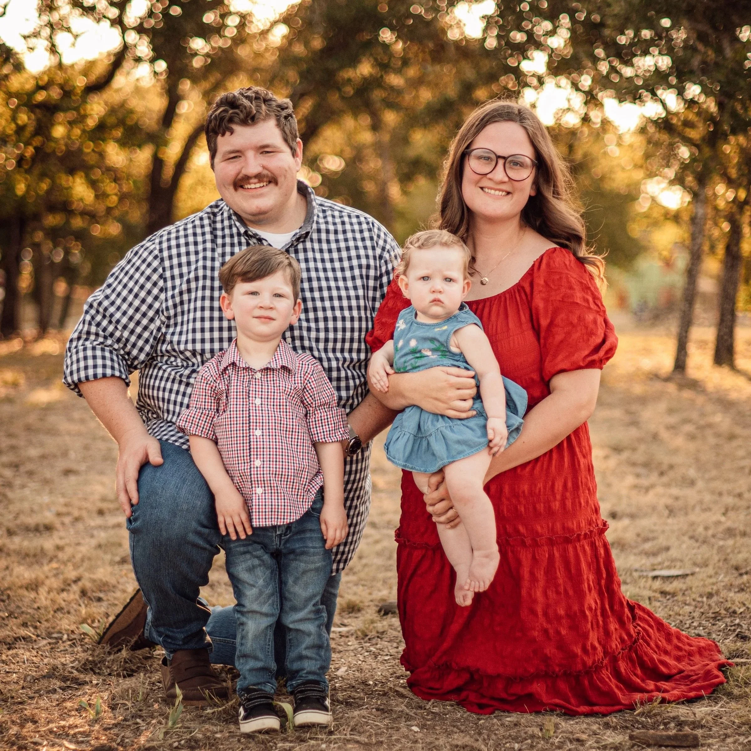 A family of four posing outdoors in a park during autumn, with trees and sunlight in the background. The father has brown curly hair and a mustache, wearing a checkered shirt. The mother has long brown hair, glasses, and is wearing a red dress. Their young son is in front, wearing a red and white checkered shirt, jeans, and sneakers. The daughter is being held by the mother, wearing a blue dress with floral patterns and has curly hair.