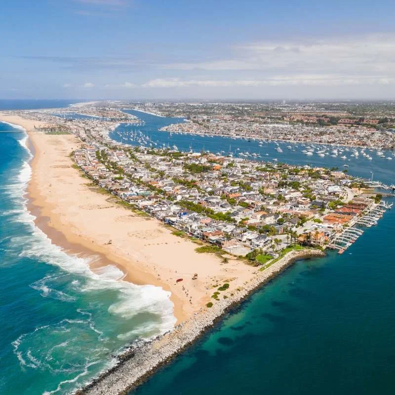 Aerial view of a coastal urban area with a sandy beach on the left, a body of water with boats in the middle, and residential neighborhoods on the right, extending into a distant horizon.