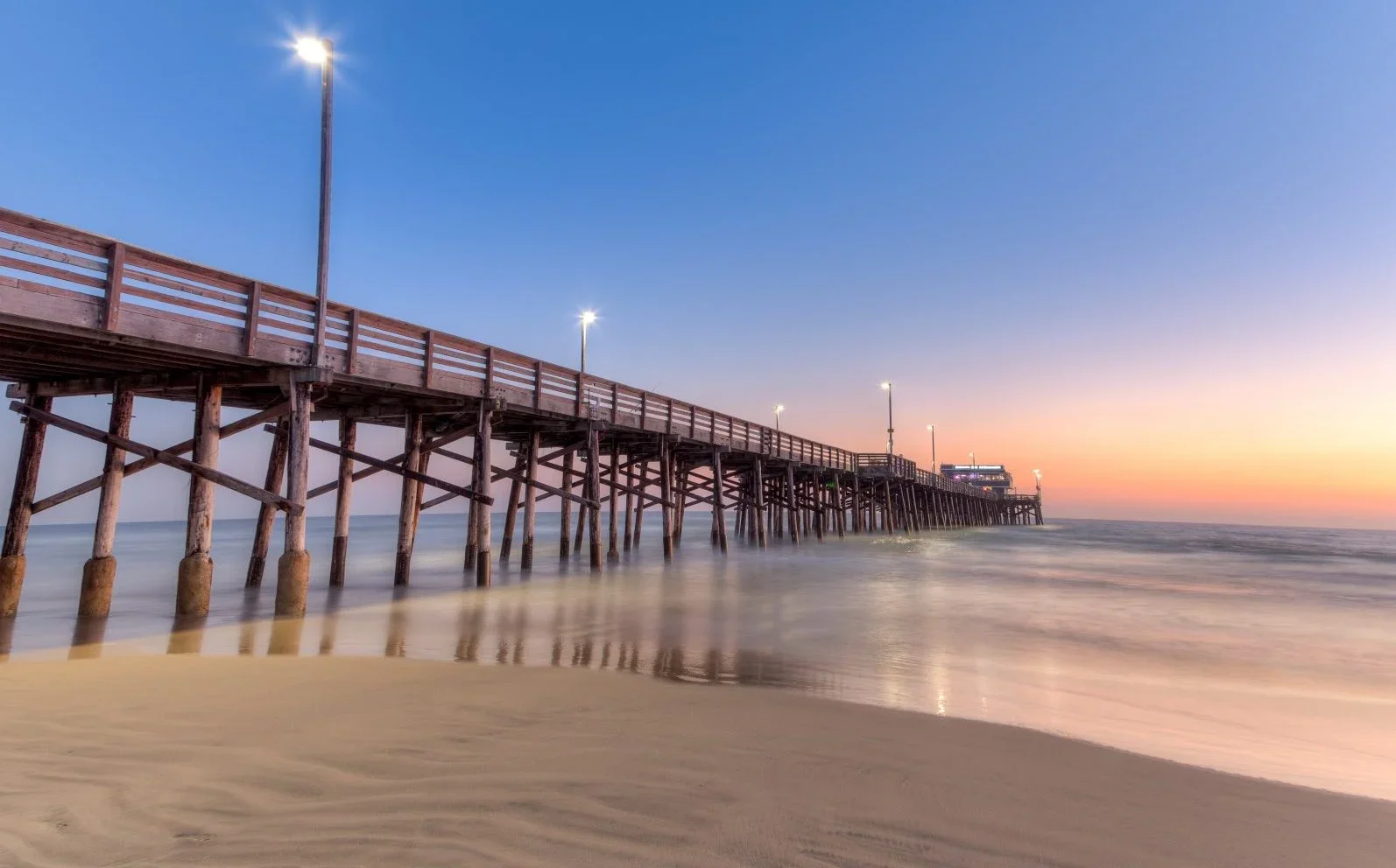 Wooden pier extending into the ocean at sunset, with a sandy beach in the foreground and lights along the pier.