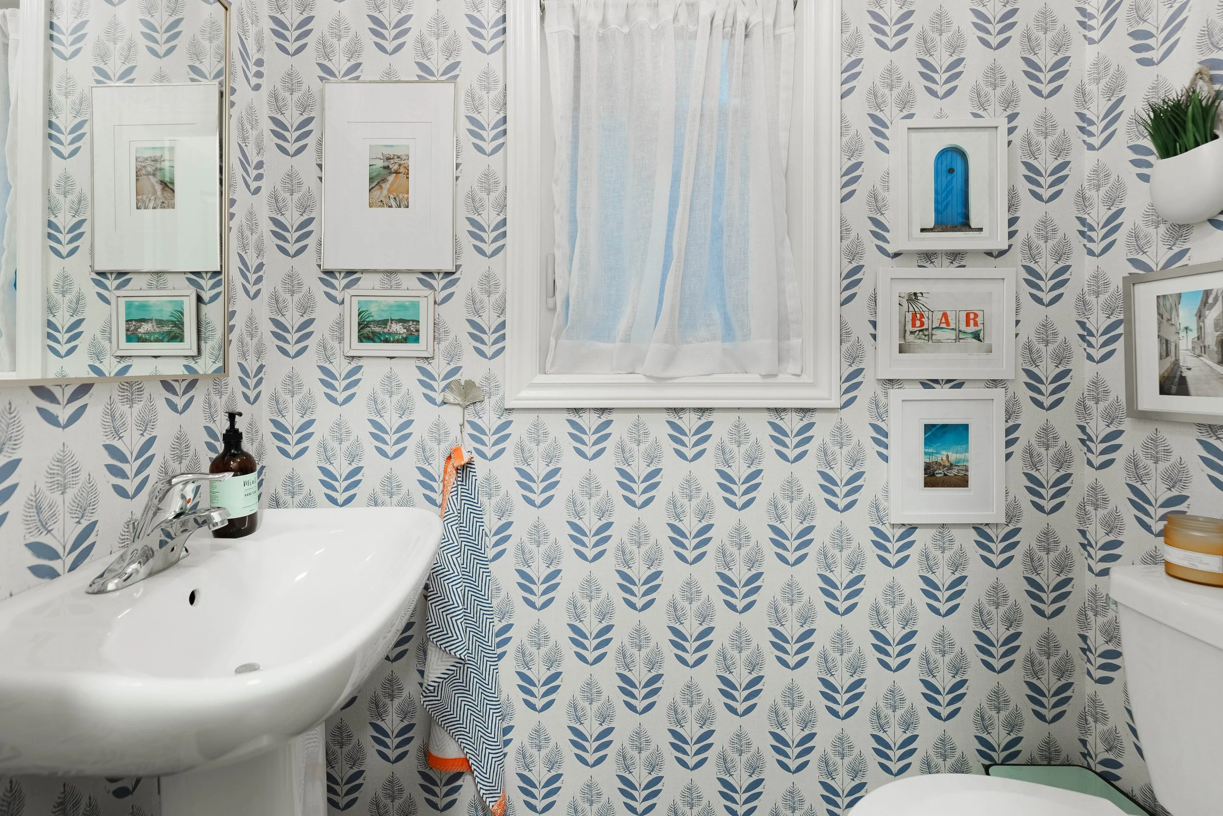 A small bathroom with blue and white patterned wallpaper and a white-framed window with sheer curtains. The wall has framed pictures, a small wall shelf with a plant, a mirror, and a towel hanging beside the sink. There is a white sink with a bottle 