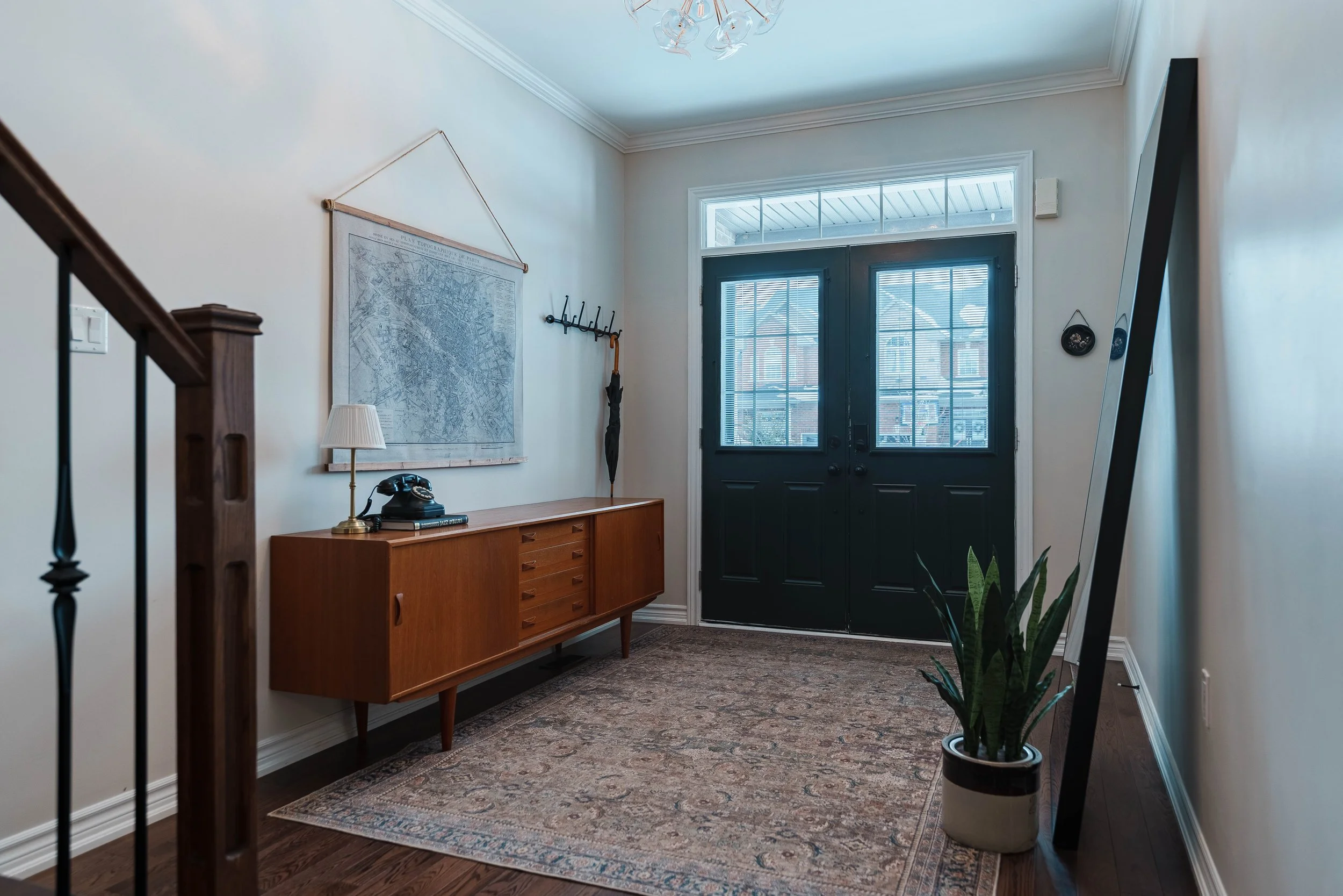 Entryway with black double doors, wooden sideboard, vintage rotary phone, small lamp, wall map, mirror, houseplant, and hooks on the wall.
