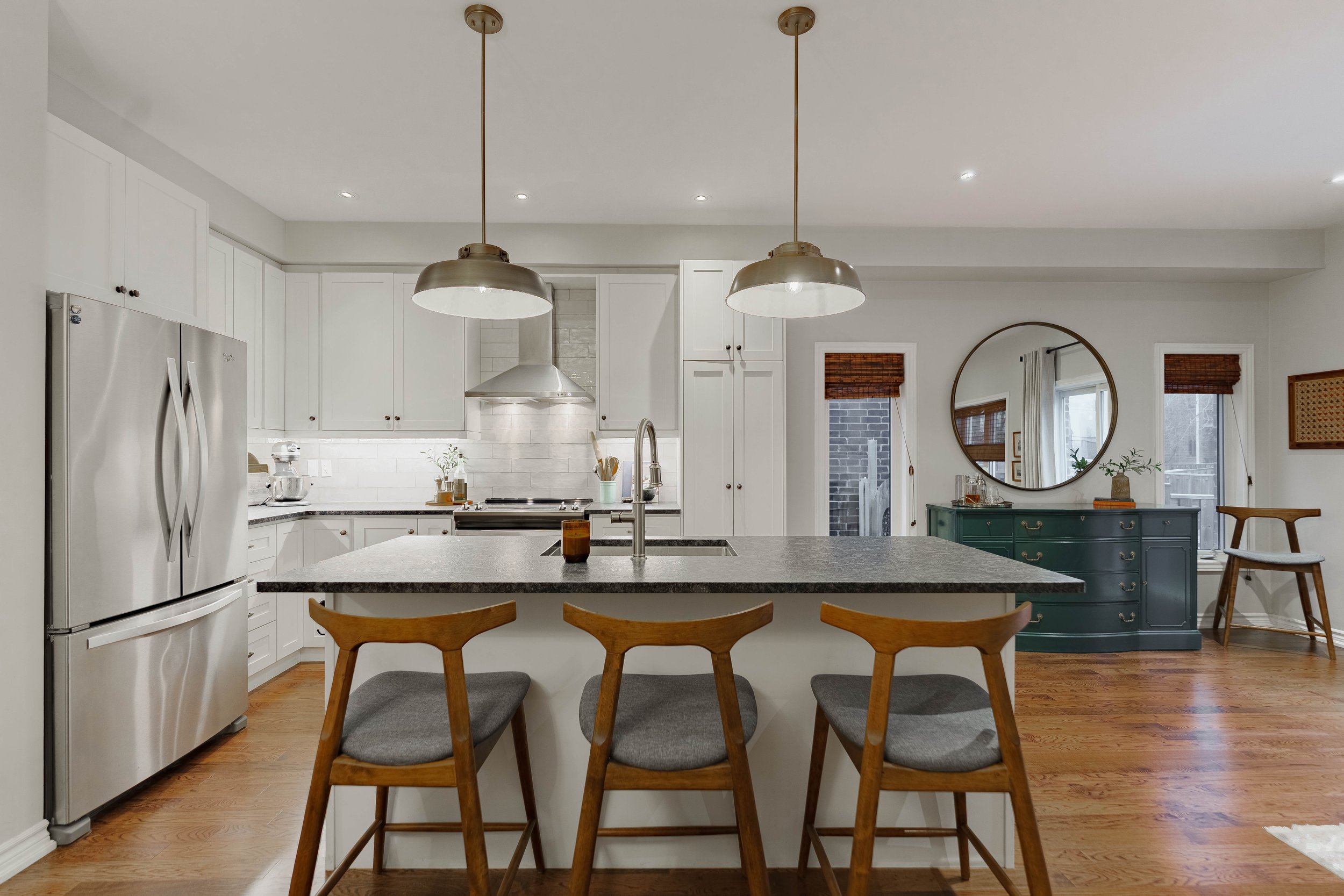 Modern kitchen with white cabinets, stainless steel refrigerator, and a central island with a sink. Two pendant lights hang above the island, and hardwood floors are visible. In the background, there is a dark green sideboard with a round mirror abov
