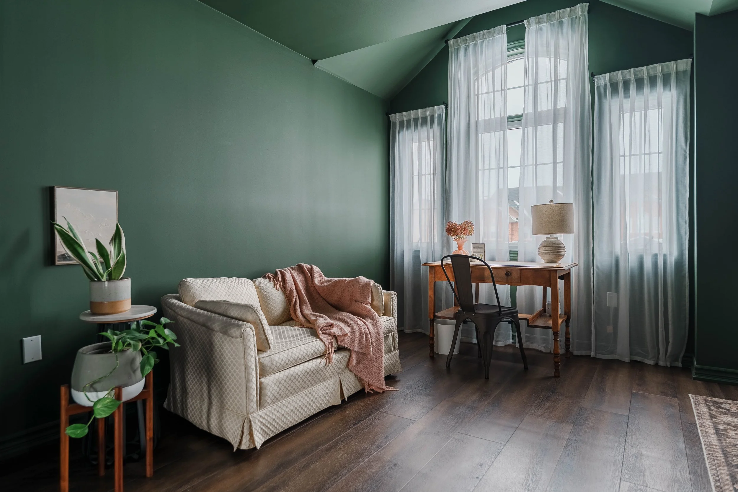 A cozy living room with green walls, a white sofa with a pink throw blanket, potted plants, wooden flooring, and a window with flowing white curtains. There's a desk with a lamp and flowers, and a black chair in front of the window.