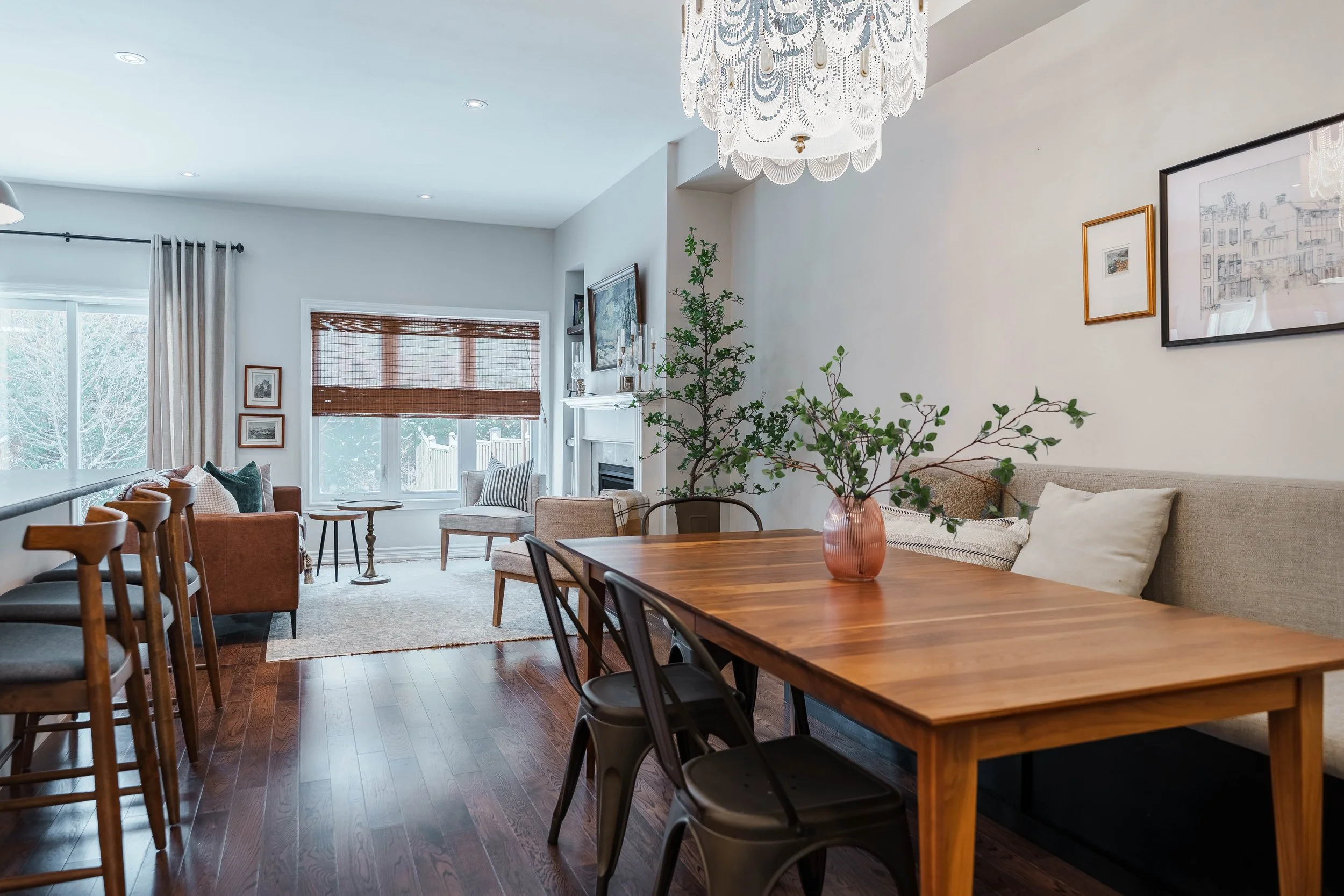 Open-concept living and dining area with hardwood floors, white walls, and large windows. The living room has a white rug, two armchairs, a small side table, and framed pictures on the wall. The dining area features a wooden table with a pink vase ho