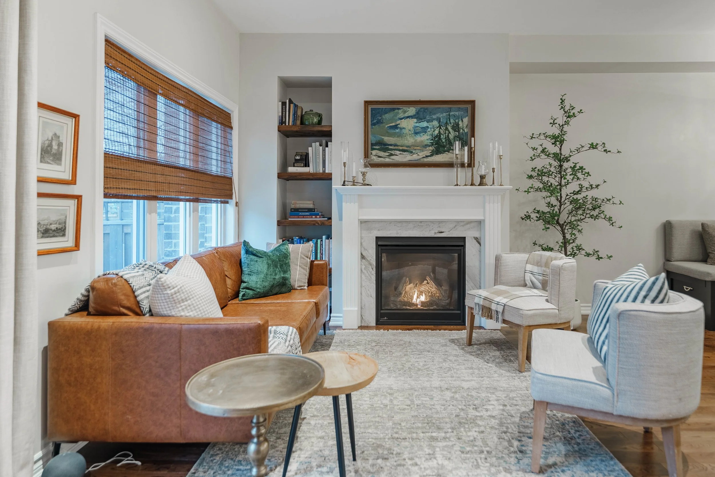 Living room with a fireplace, brown leather sofa with pillows, two white armchairs, window with wooden blinds, bookshelf, artwork above fireplace, and a potted plant.