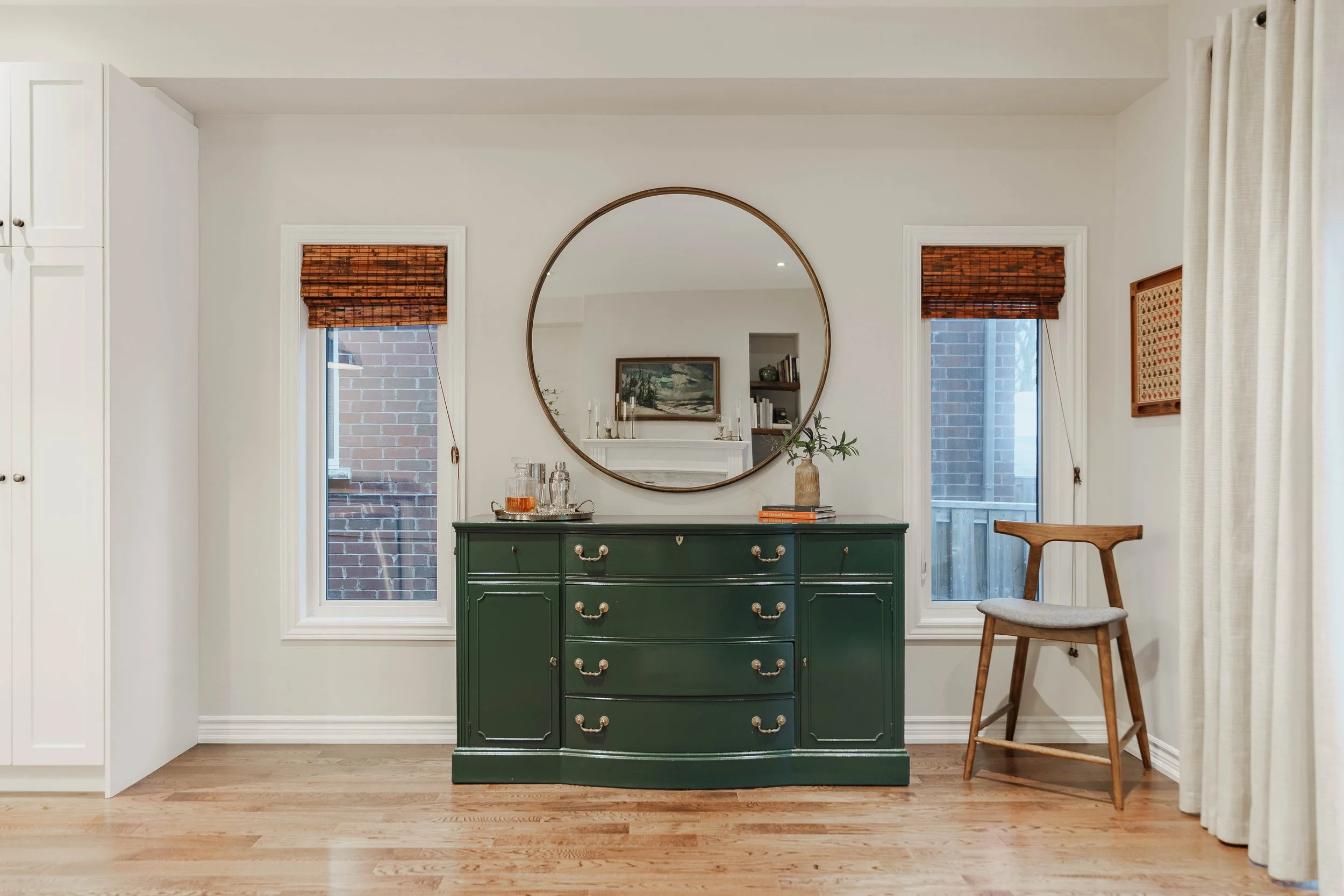 A green dresser with brass handles in a room, topped with a tray of drinks, a vase with green plants, and books. A large round mirror hangs above it. Two windows with bamboo blinds are on either side, and a wooden chair is on the right.