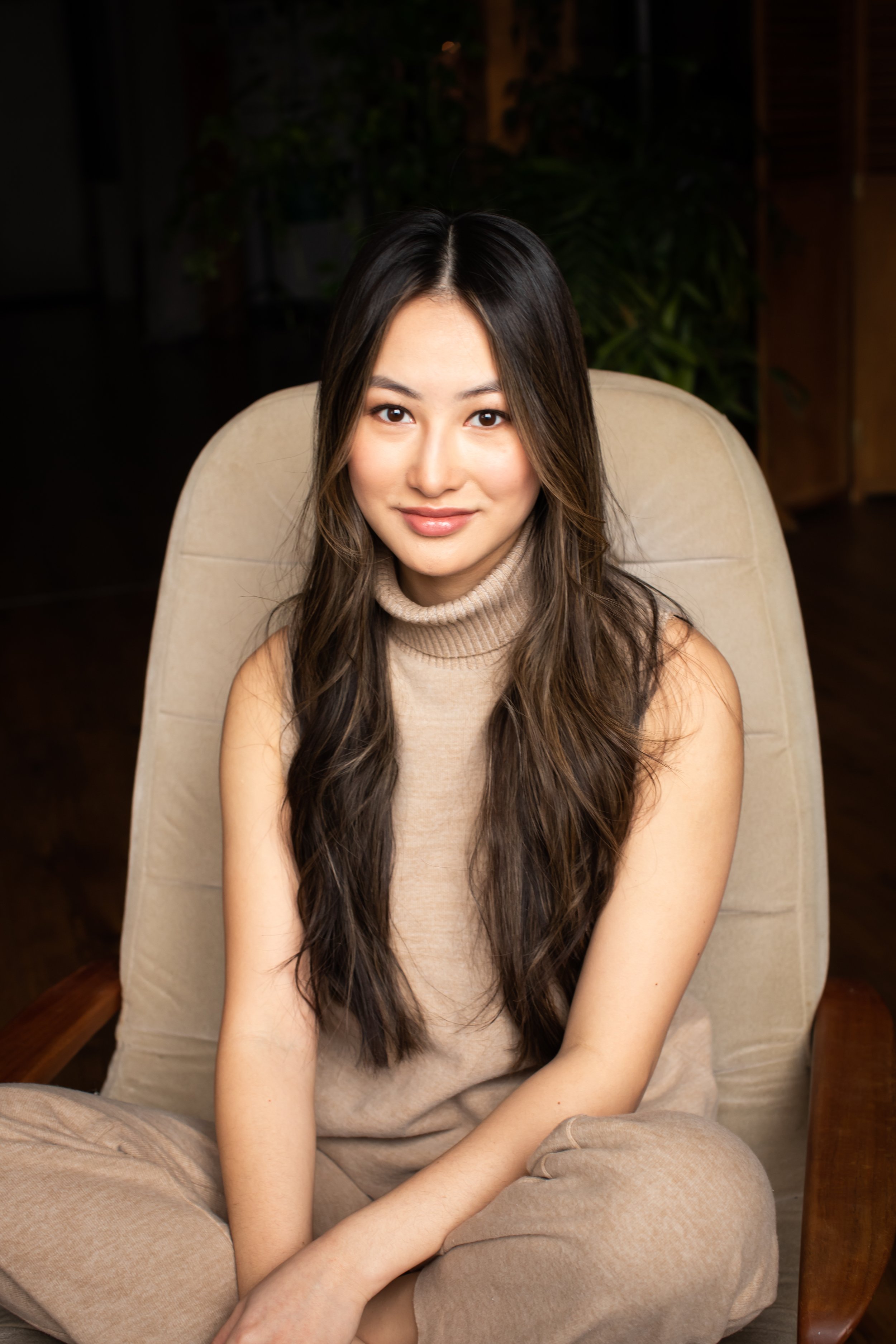 A young woman with long dark wavy hair and light skin, wearing a beige sleeveless turtleneck top, smiling and sitting cross-legged on a light-colored armchair in an indoor setting.