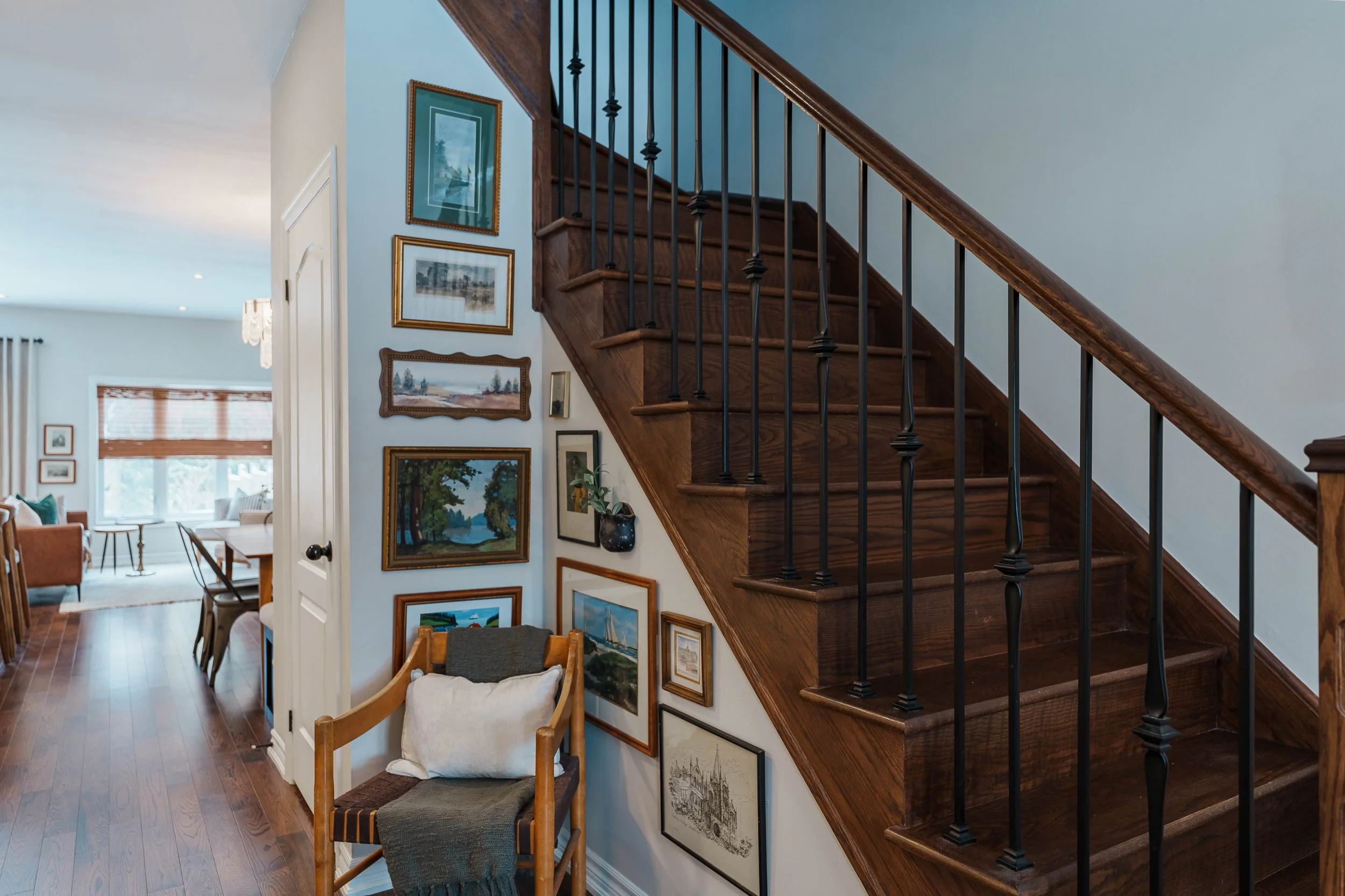 Living room with wooden flooring, a gallery wall with framed artworks, a wooden staircase with black metal railings, and a sitting area with armchairs and windows in the background.