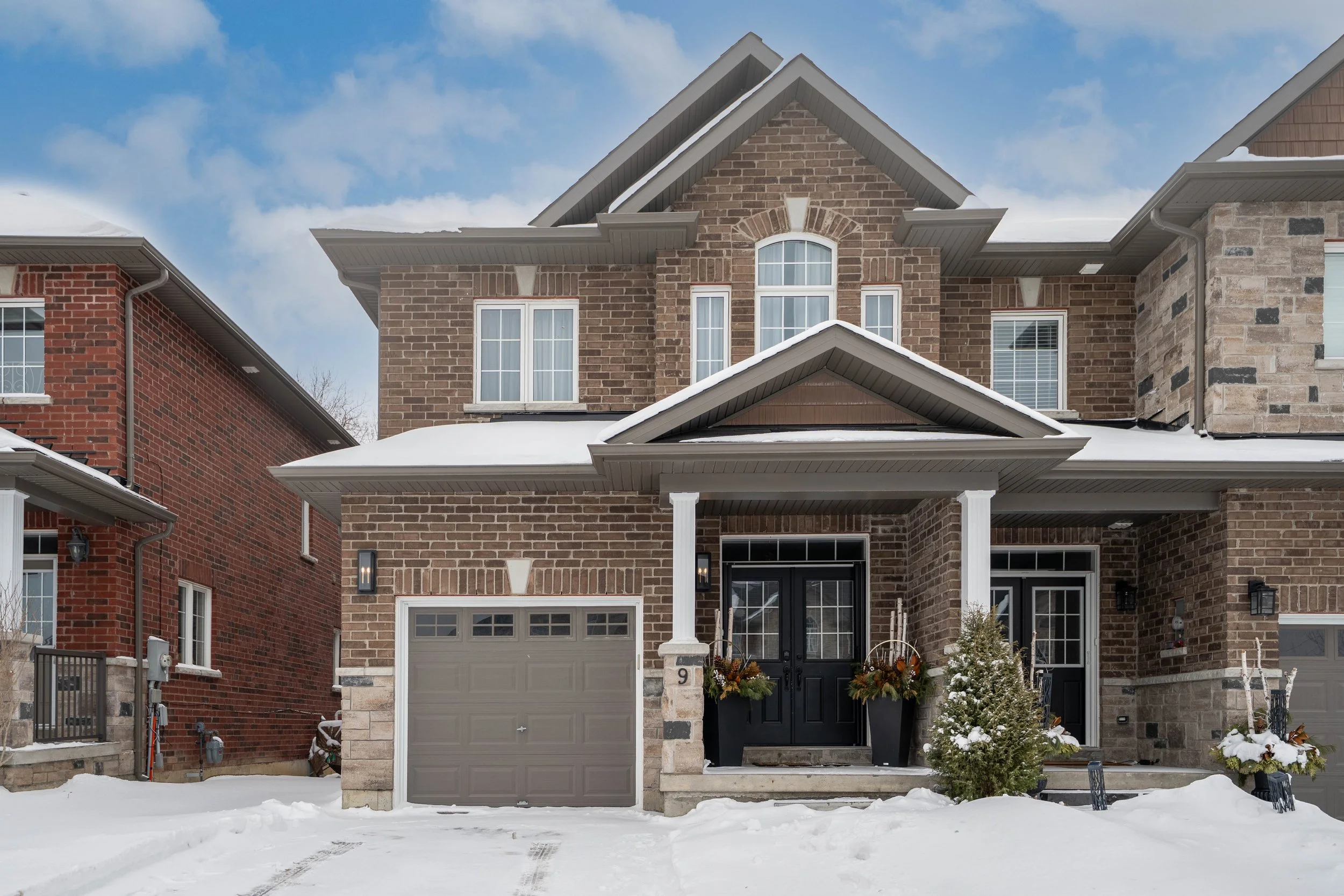 A large two-story brick house with an attached garage, snow-covered driveway, and winter decorations outside, including potted plants and small snow-covered trees.
