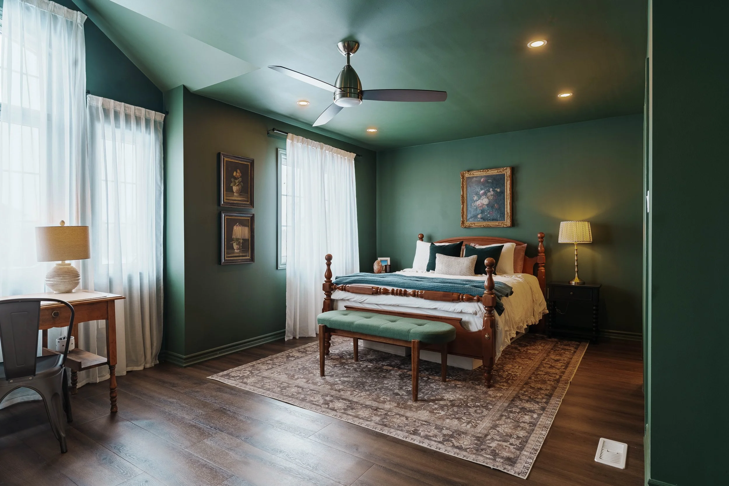 A cozy bedroom with a green color scheme, featuring a wooden bed with comfortable pillows, a bench at the foot of the bed, nightstands with lamps, window curtains, a ceiling fan, and framed artwork on the walls.