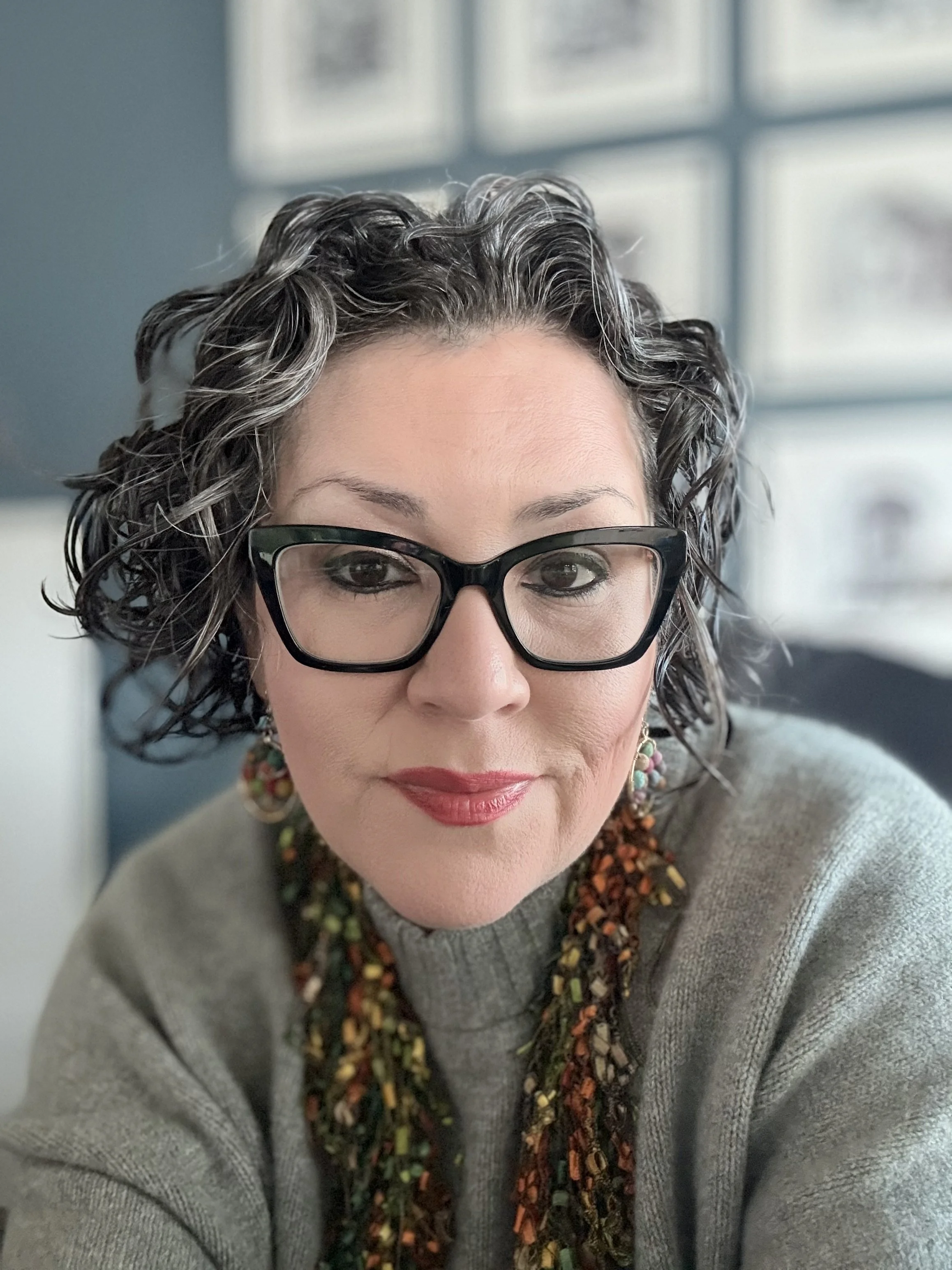 Retrato de una mujer con cabello rizado gris y oscuro, gafas negras, camiseta gris y bufanda colorida, en un ambiente con ventanas en el fondo.