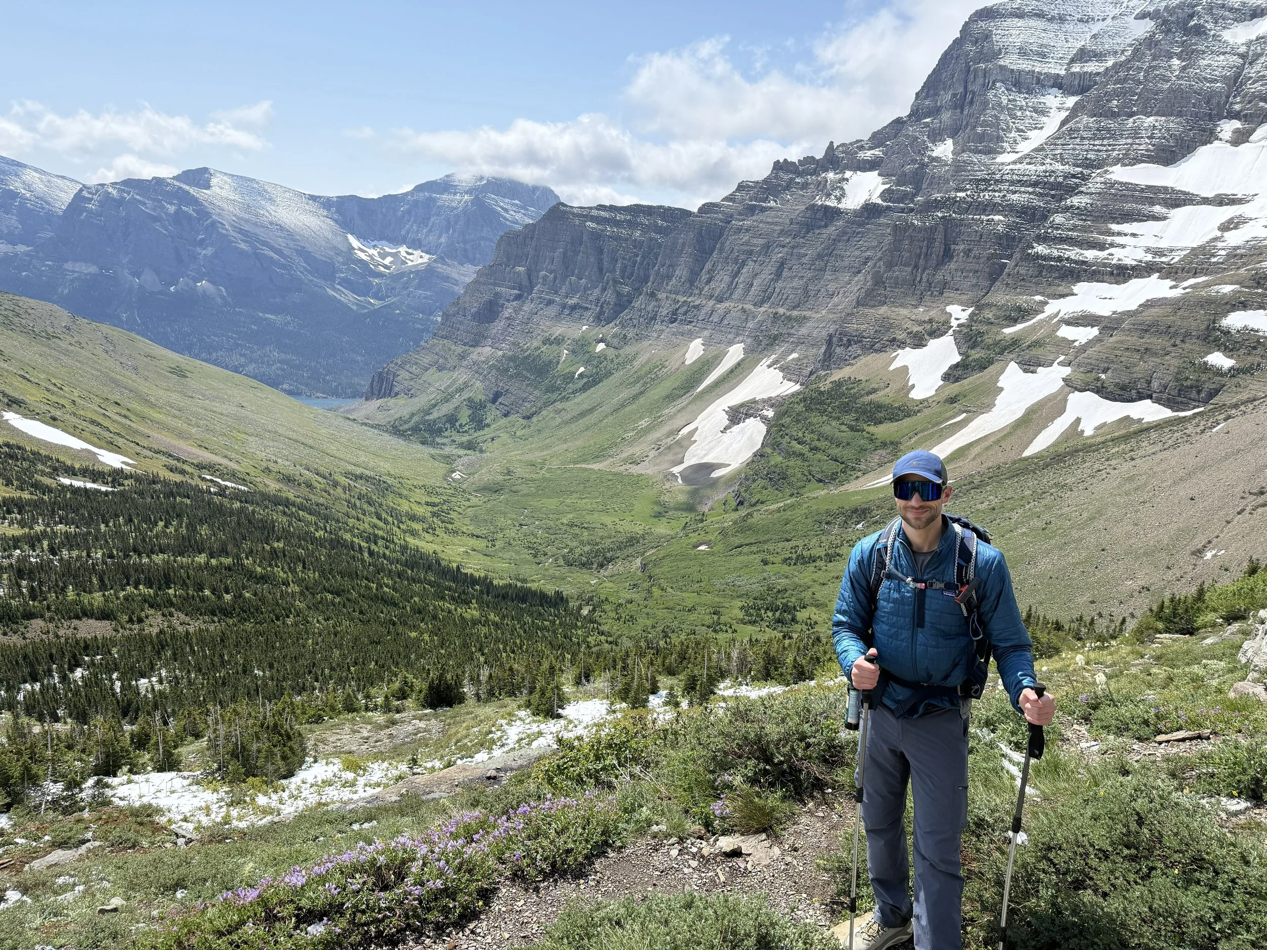 A man hiking in a mountainous area with snow-capped peaks, green forests, and a trail, holding poles and wearing a backpack, sunglasses, and outdoor gear.