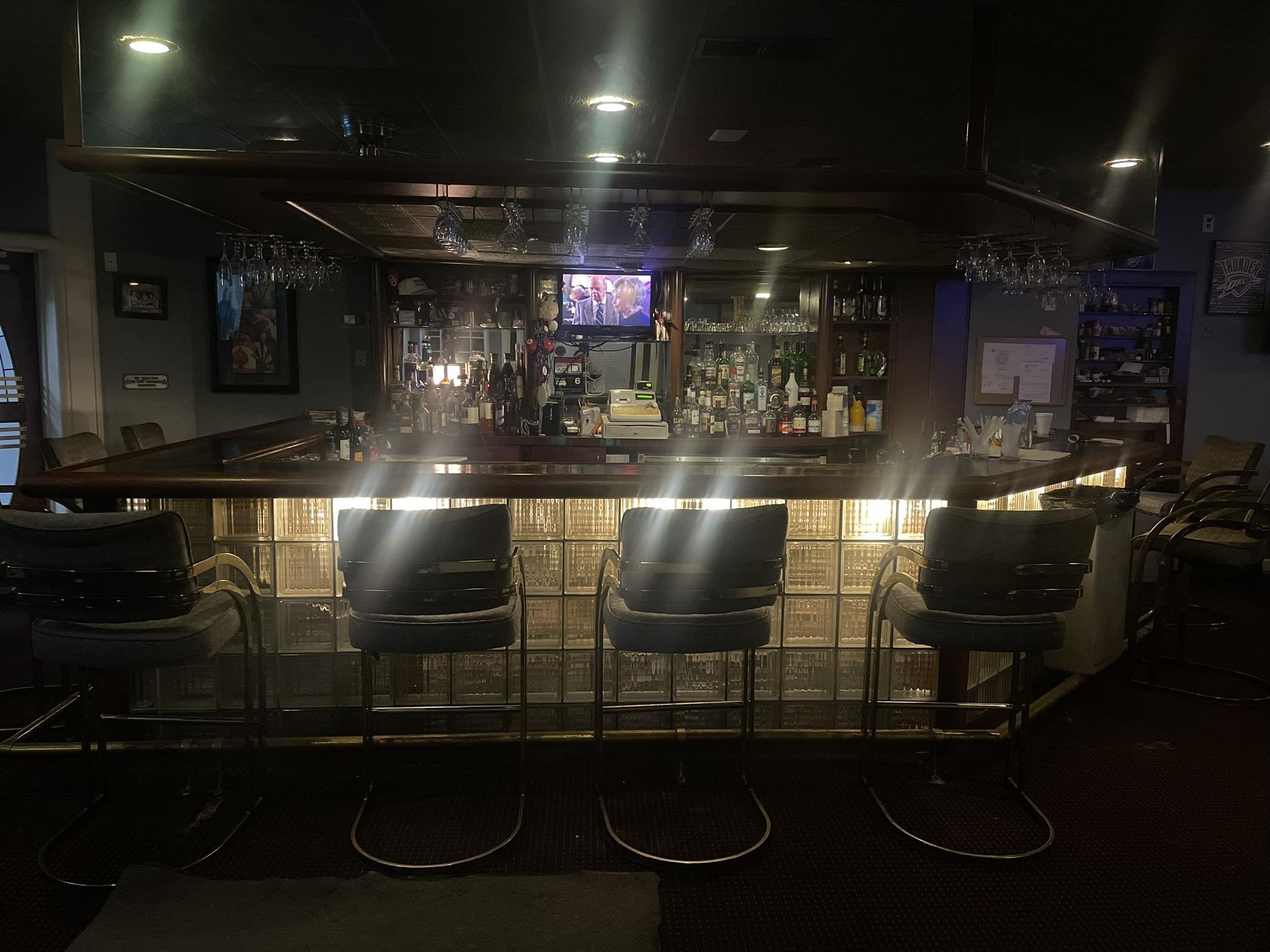 Empty bar with four barstools, illuminated case with bottles of alcohol, and TV on the wall in a dimly lit sports bar.