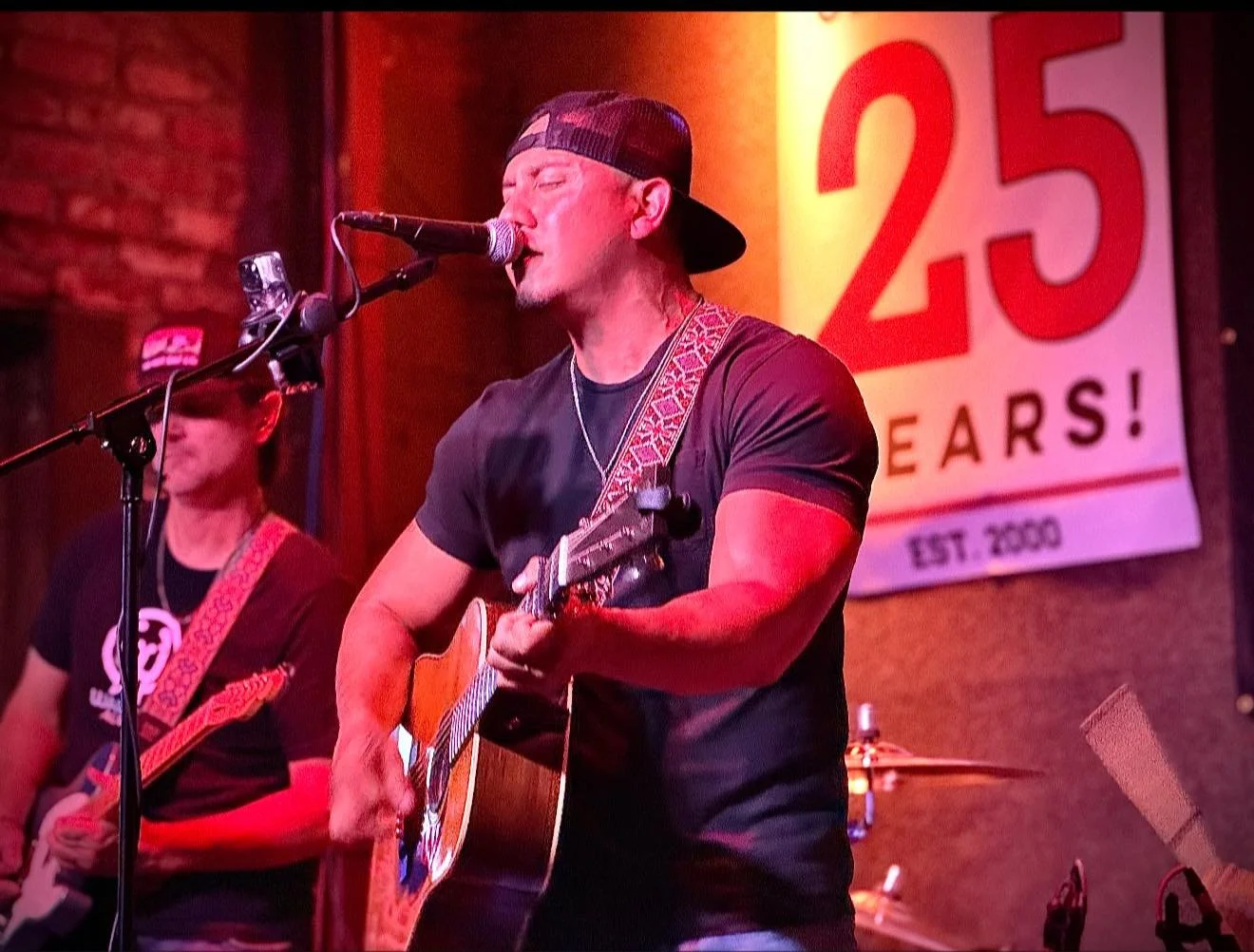 singing into a microphone and playing guitar on stage during a live performance. He wears a black cap backward and a black t-shirt, with a patterned guitar strap. There is another musician in the background, also playing guitar. The stage is lit with pink and orange lighting, and a sign celebrating 25 years since 2000 is visible in the background.