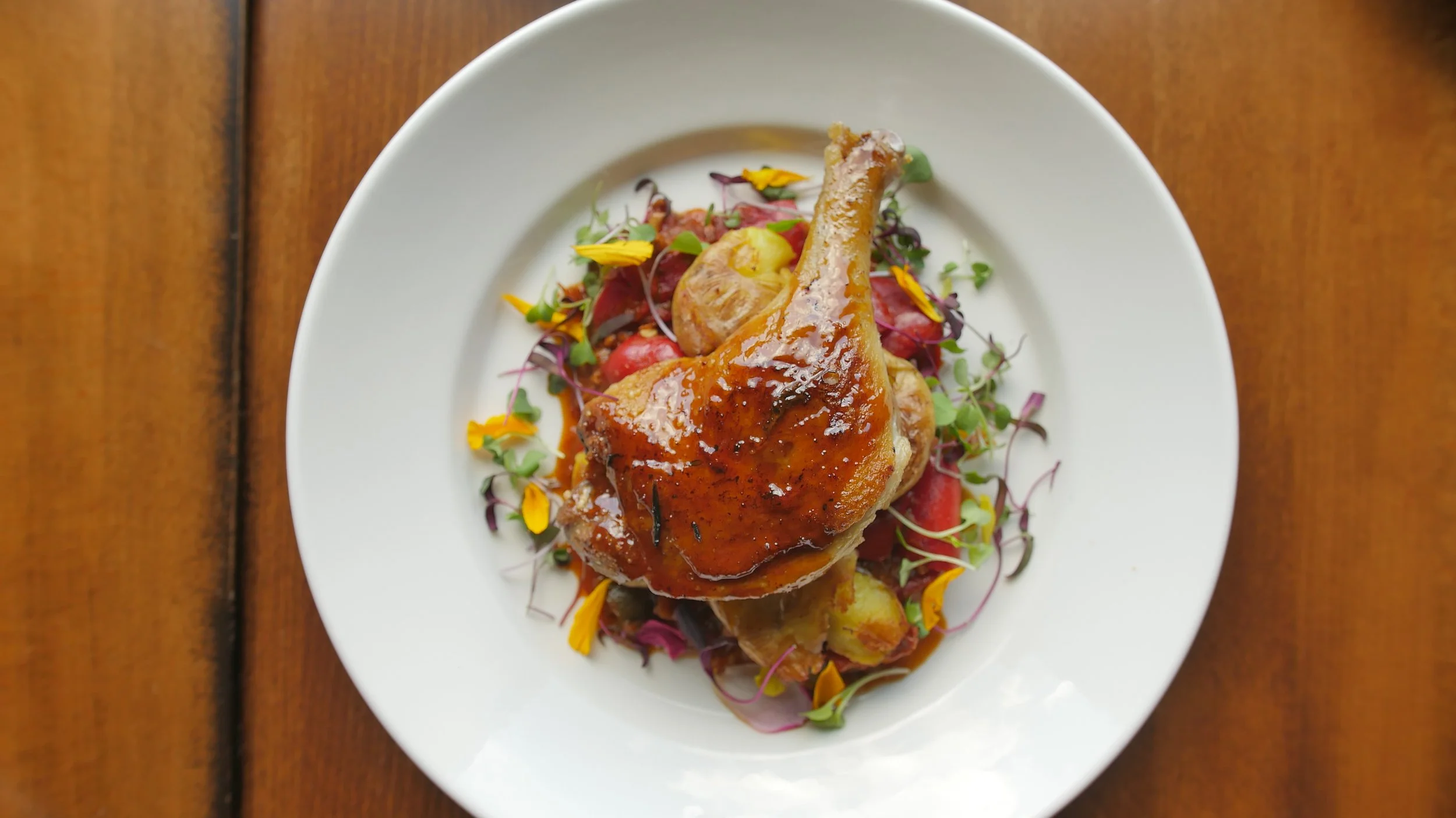 A white plate with a roasted chicken leg glazed with sauce, served on a bed of mixed greens, cherry tomatoes, and garnished with edible yellow and purple flowers, on a wooden table.