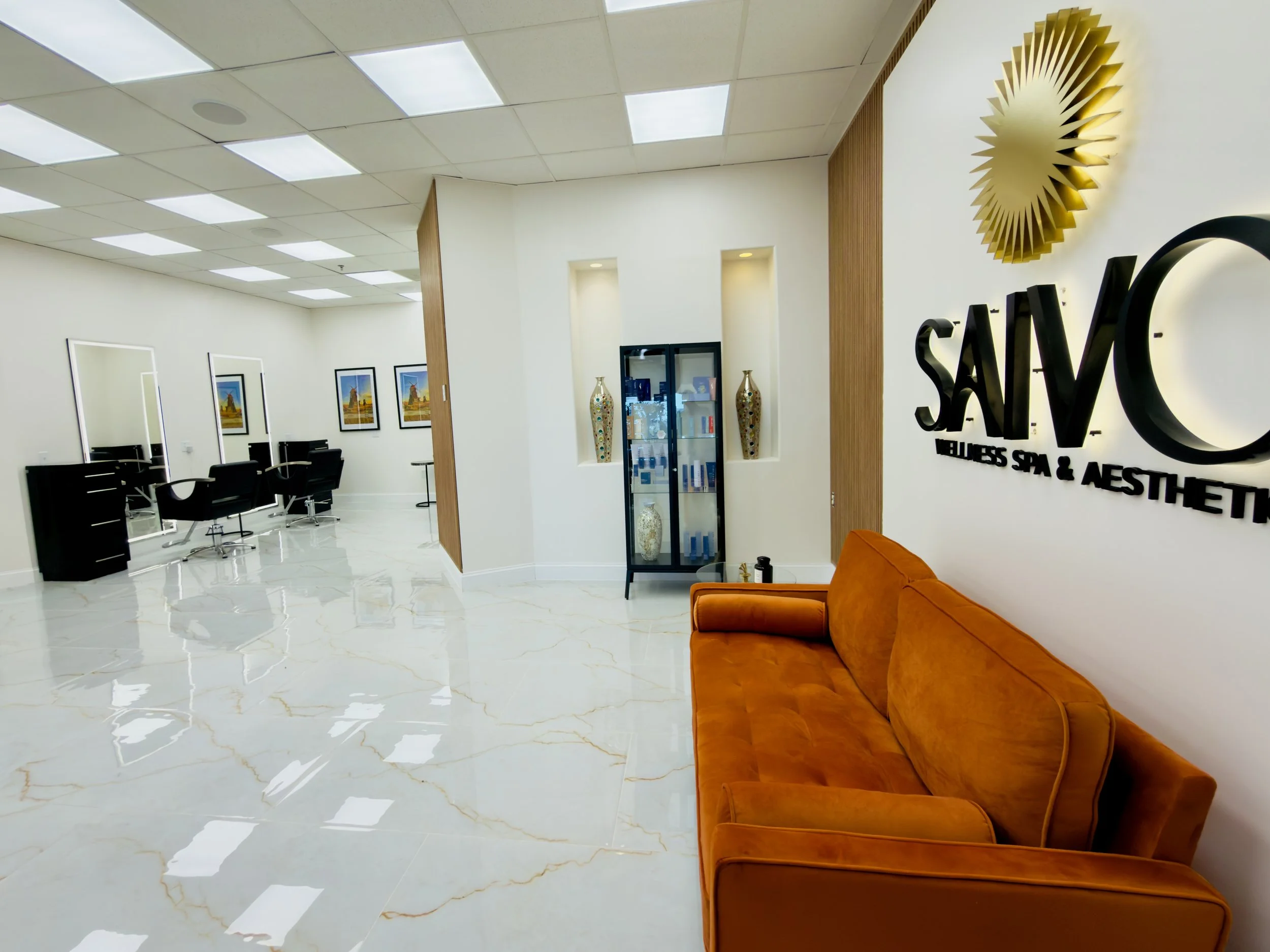 Reception area of a wellness salon with white walls, paintings, a velvet orange sofa, and signage that reads 'Salon Wellness Spa & Aesthetics'.