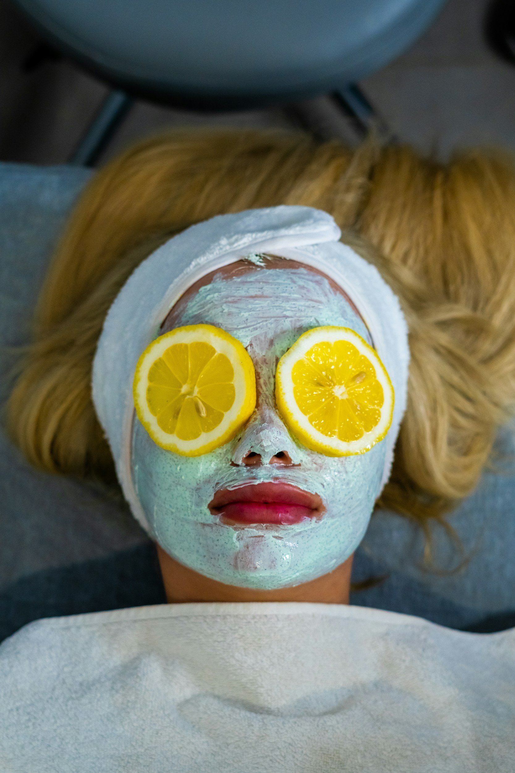 A person lying down with a facial mask applied, two lemon slices are placed over their eyes, and they are wrapped in a white towel around their head with blonde hair visible.