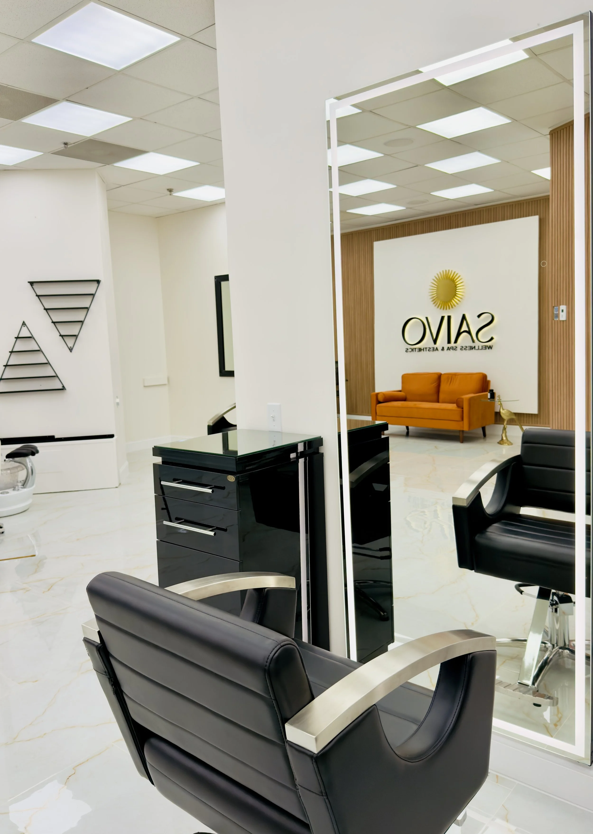 Modern salon interior with black leather chairs, a small black rolling cabinet, marble flooring, and a wall with the salon's logo and orange sofa.