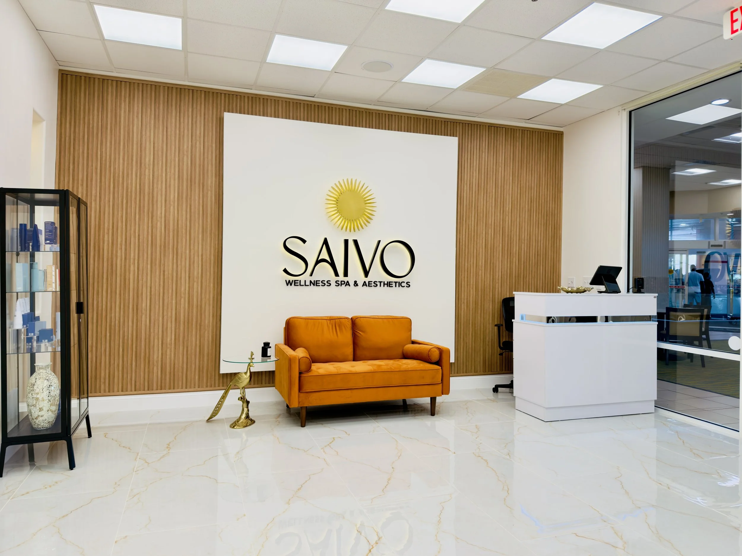 Interior of wellness spa and aesthetics reception area with a wooden accent wall, white signage with a yellow sun logo and the name 'SAVO'. Orange sofa, glass side table, decorative items, and a reception desk with a tablet. Large window showing an o