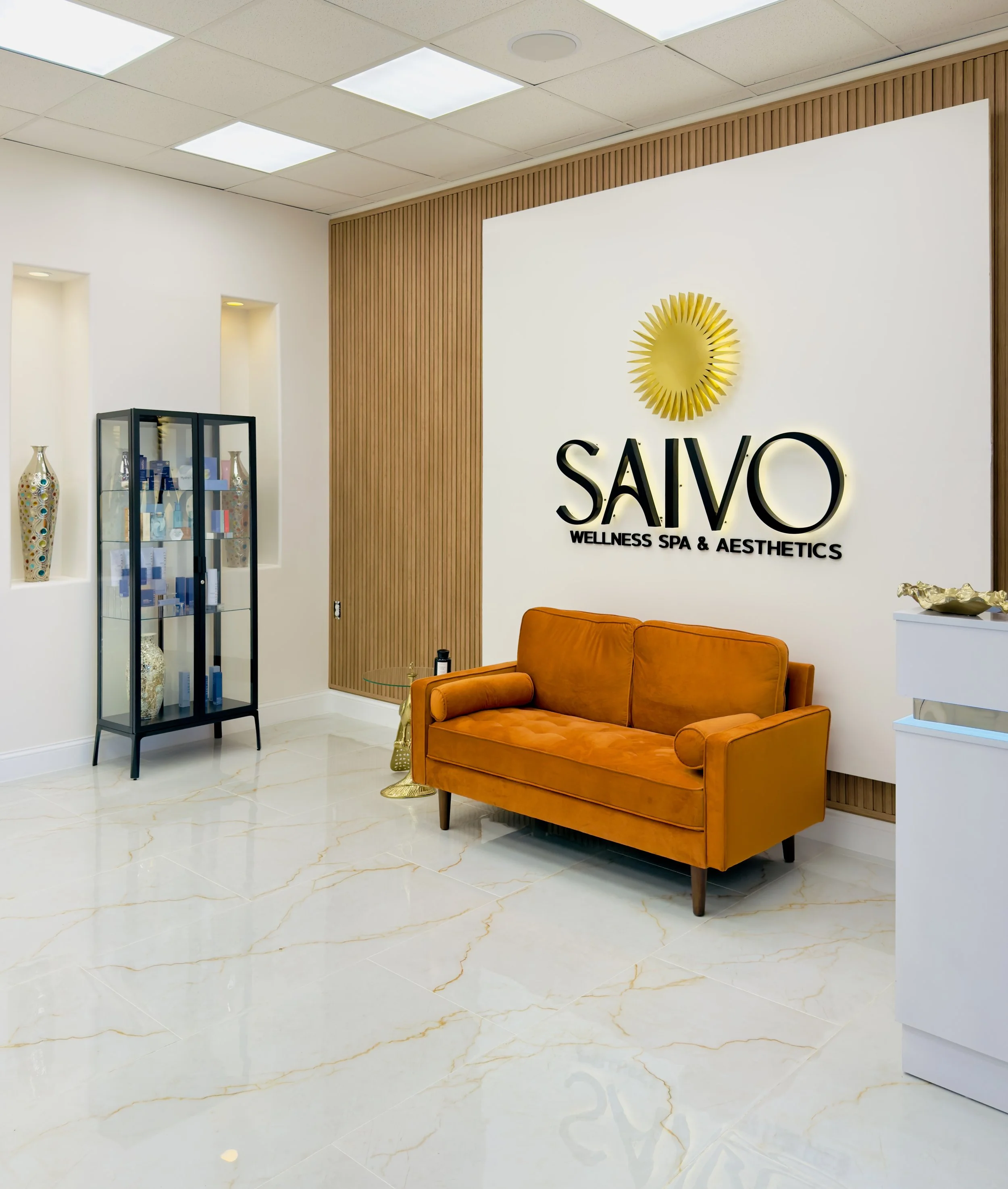 Interior of a wellness spa with a reception area. The wall has a sign that reads 'SAIVO Wellness Spa & Aesthetics' with a sunburst design. There is an orange sofa, a glass side table, a display cabinet with spa products, and decorative vases. The flo