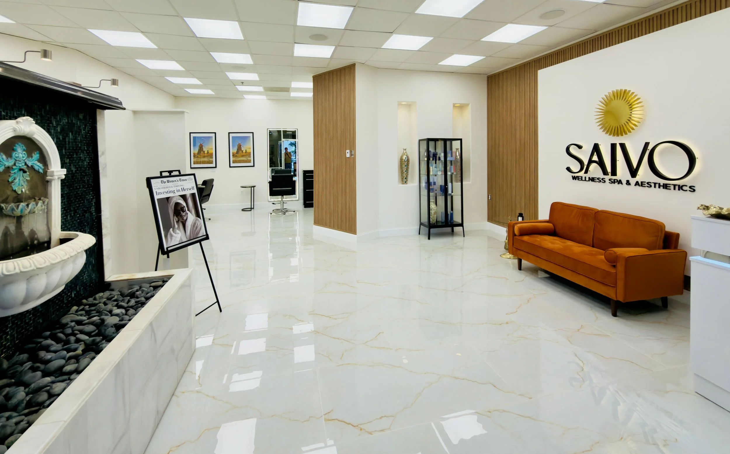 Lobby of a wellness spa and aesthetics center with white walls, wooden accents, and a tan sofa. Features include artwork, a display case with decorative items, a fountain with black pebbles, and signage for 'SAVO Wellness Spa & Aesthetics' on the wal