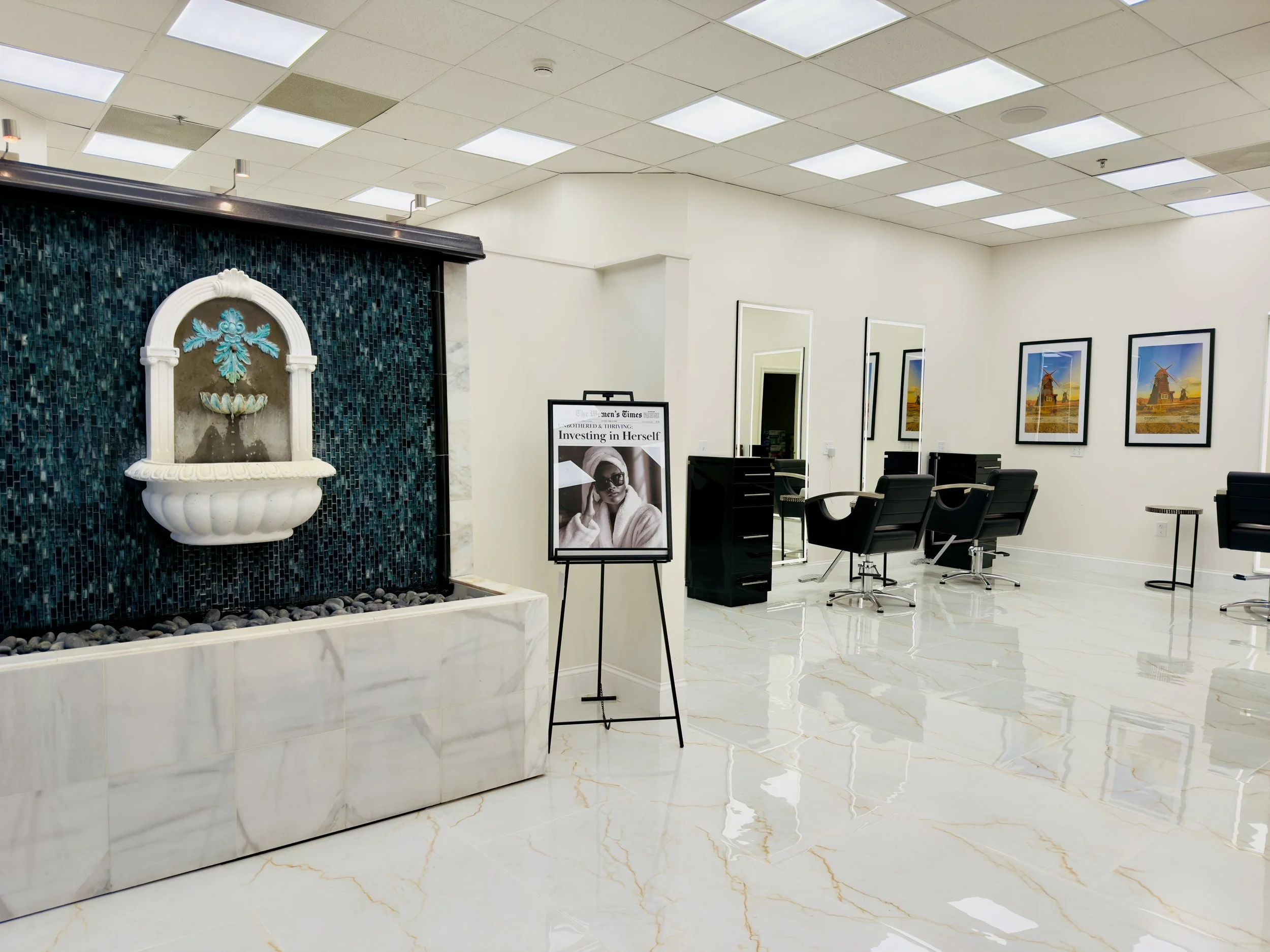Interior view of a salon or spa, with a white and blue fountain on the left, framed pictures of windmills on the wall, black chairs, and large mirrors, all with bright ceiling lighting and a marble floor.
