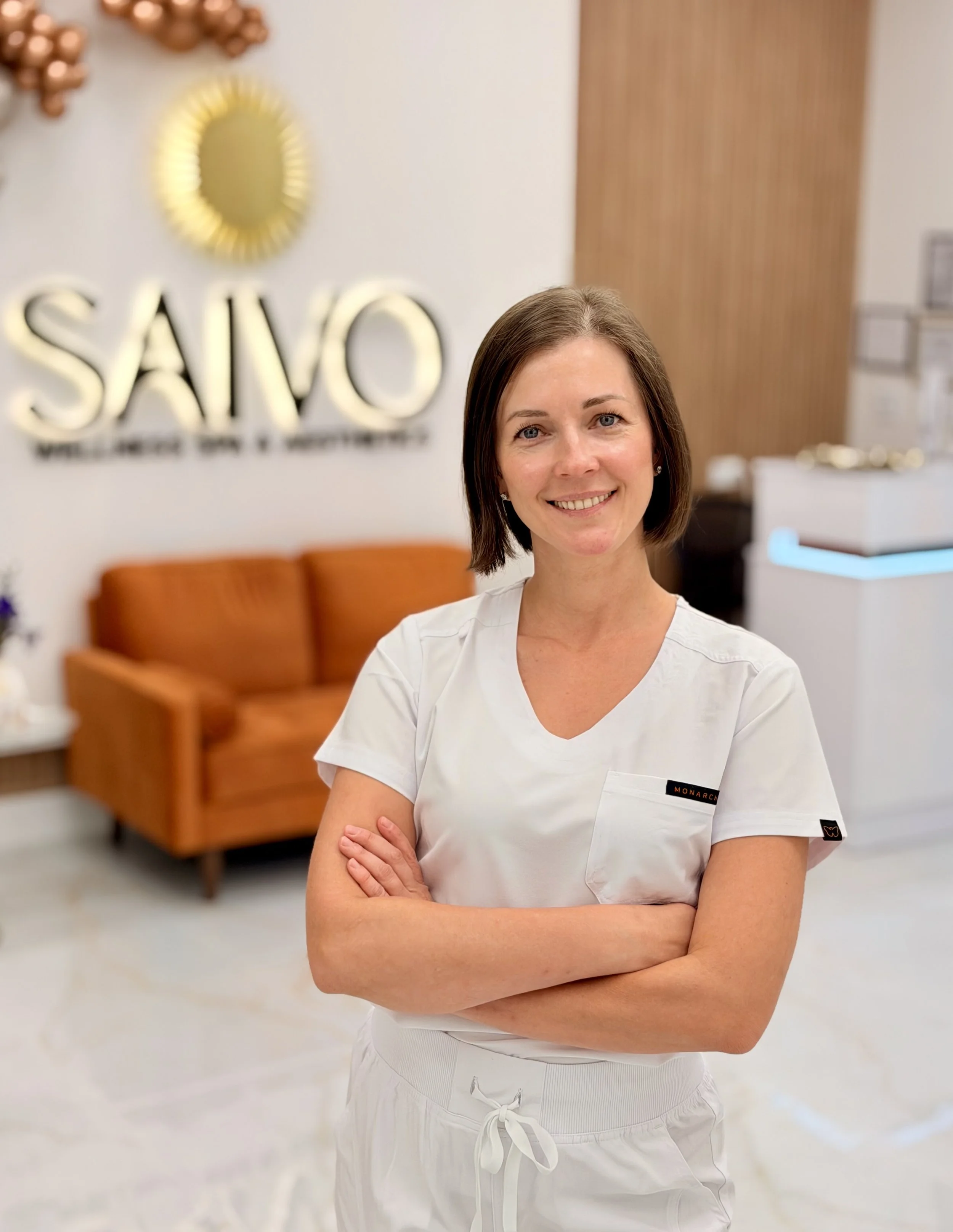 Smiling woman in white medical scrubs with arms crossed inside a medical facility, with a sign reading 'SAVO' in the background.