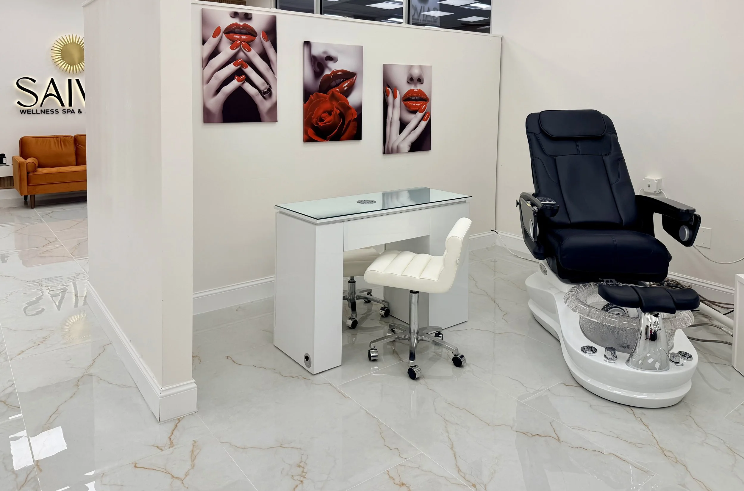 A modern beauty treatment room with a black facial chair and white manicure table with chairs, decorated with vibrant pink, red, and orange lipstick-themed artwork on the walls. The room has white marble flooring and is part of a wellness spa.