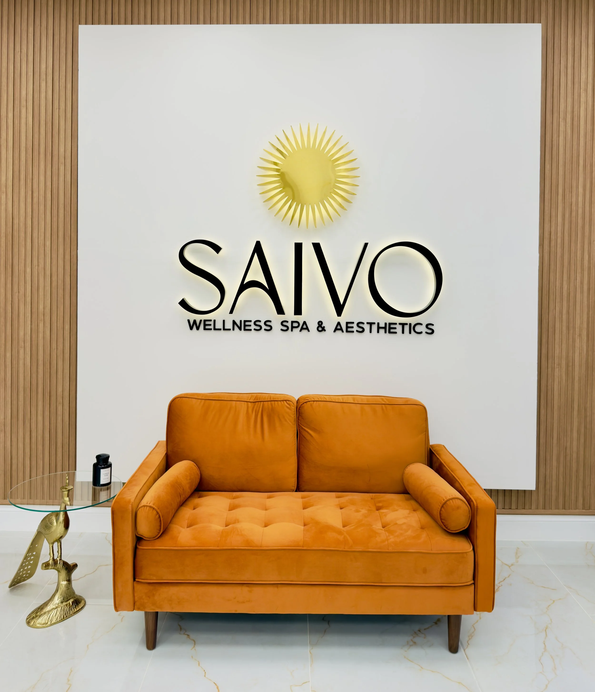 A brown vintage-style sofa with rounded arms and dark wooden legs, placed in front of a white wall with a logo for 'SAVO Wellness Spa & Aesthetics' featuring a gold sunburst symbol, a small round glass side table with a gold unicorn sculpture, black 