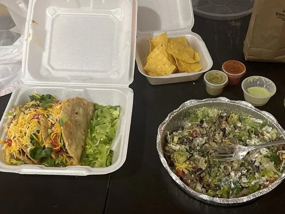 Takeout container with shredded Mexican cheese, lettuce, and a taco with a meat filling. Next to it, a foil tray of chopped salad with lettuce, vegetables, and dressing. Outside, there are small containers of salsa, guacamole, and sour cream, along w