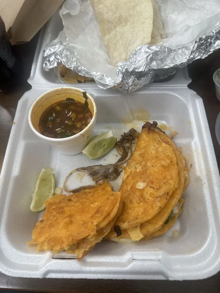 Two tacos with fried shells, filled with shredded meat and cheese, served with lime wedges, a small cup of sauce, and a piece of roasted meat on a foam tray. There are also two tortillas wrapped in aluminum foil.