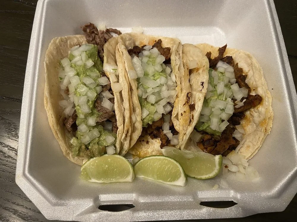 Three tacos with diced onions and chopped green peppers, served with lime wedges in a foam takeout container.