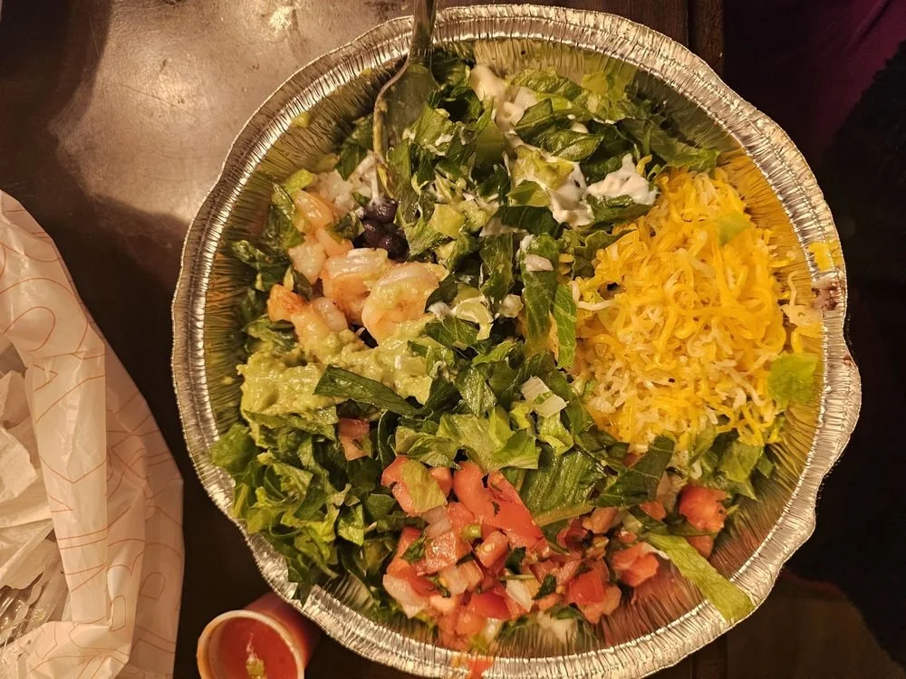 Mixed salad with shredded cheddar cheese, chopped tomatoes, lettuce, onions, and possibly shrimp in a foil container.