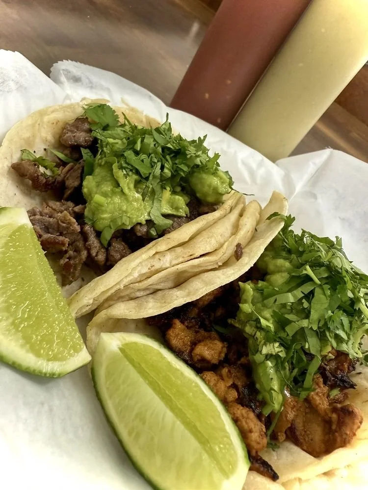 Two tacos with chopped cilantro, chopped lettuce, and grilled meat, served with lime wedges, in a paper tray with sauces in bottles in the background.